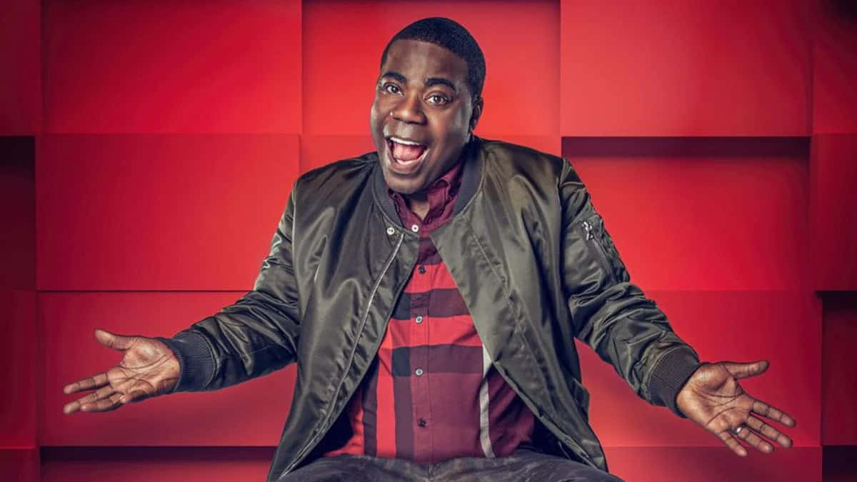 Tracy Morgan 桌布 壁紙 — 一個快樂的男人穿著飛行員夾克和格子襯衫，張開雙臂坐在大膽的紅色幾何牆前。