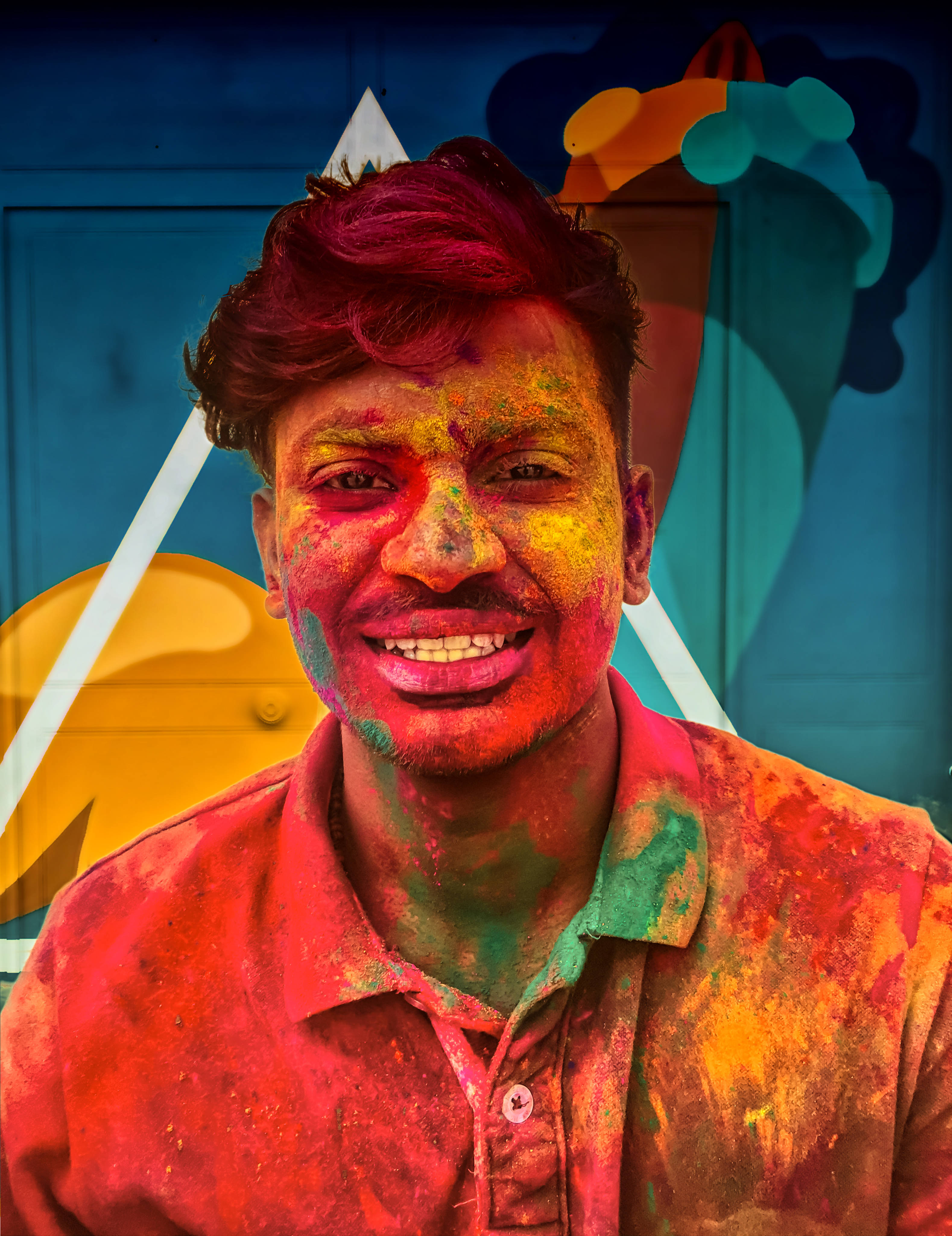 Holi 桌布 壁紙 — 一個快樂的男人臉上和襯衫上沾滿鮮豔的彩色粉末，在風格化的藍色背景前燦爛微笑。
