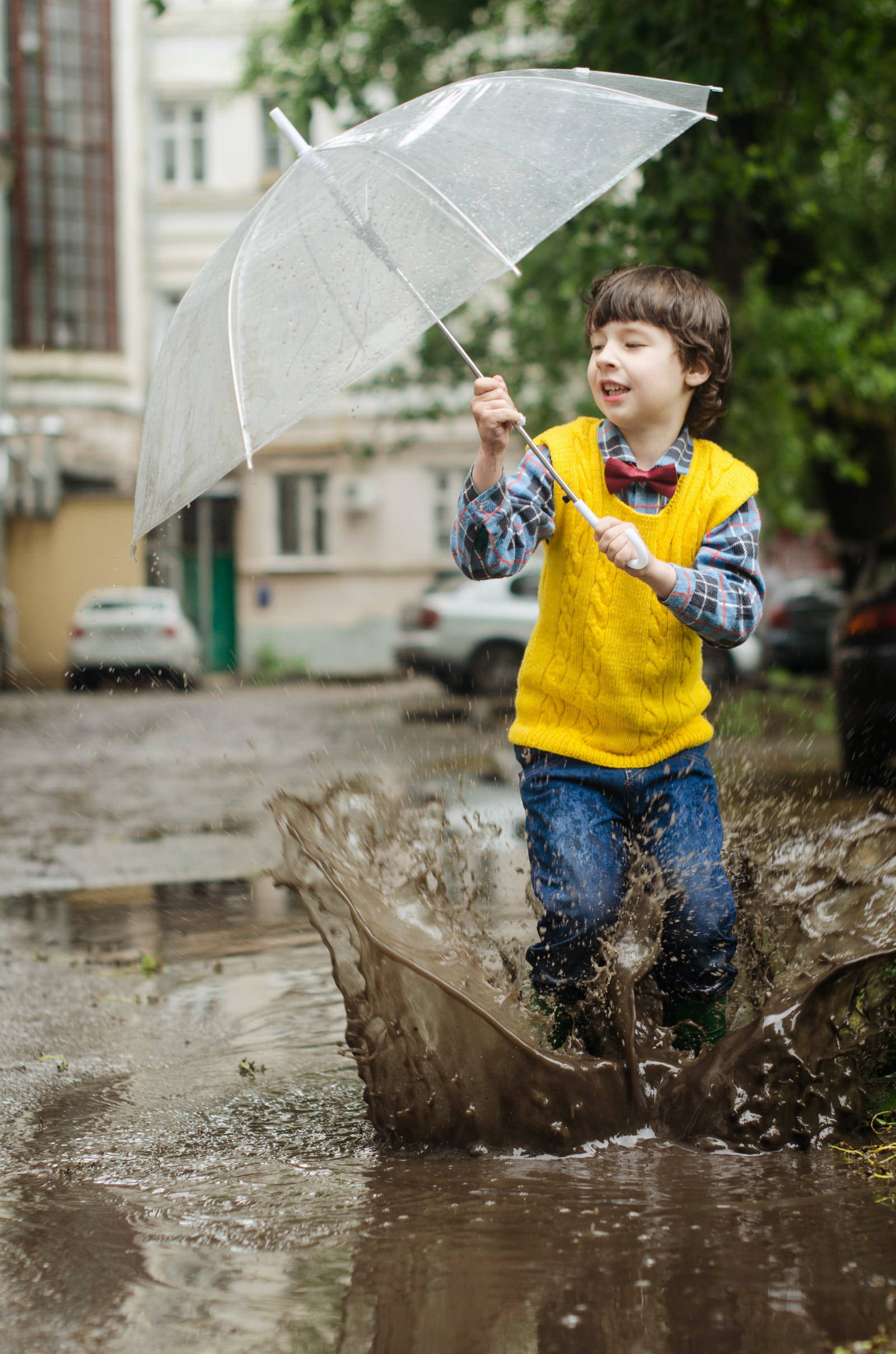 Raining 桌布 壁紙 — 一個快樂的男孩穿著黃色背心，在雨天的城市街道上拿著透明雨傘跳過泥濘的水坑。