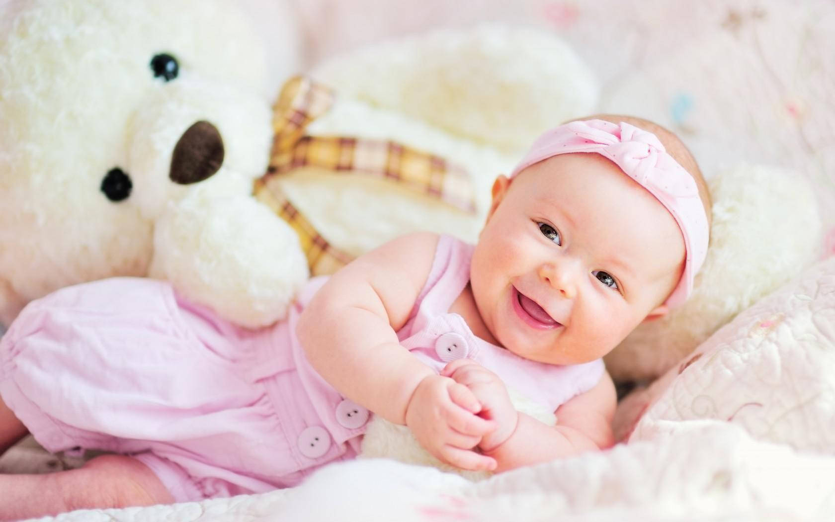Newborn 桌布 壁紙 — 一個快樂的粉紅色衣服女嬰躺在柔軟的床上，旁邊有一隻大白熊玩偶，正在大笑。