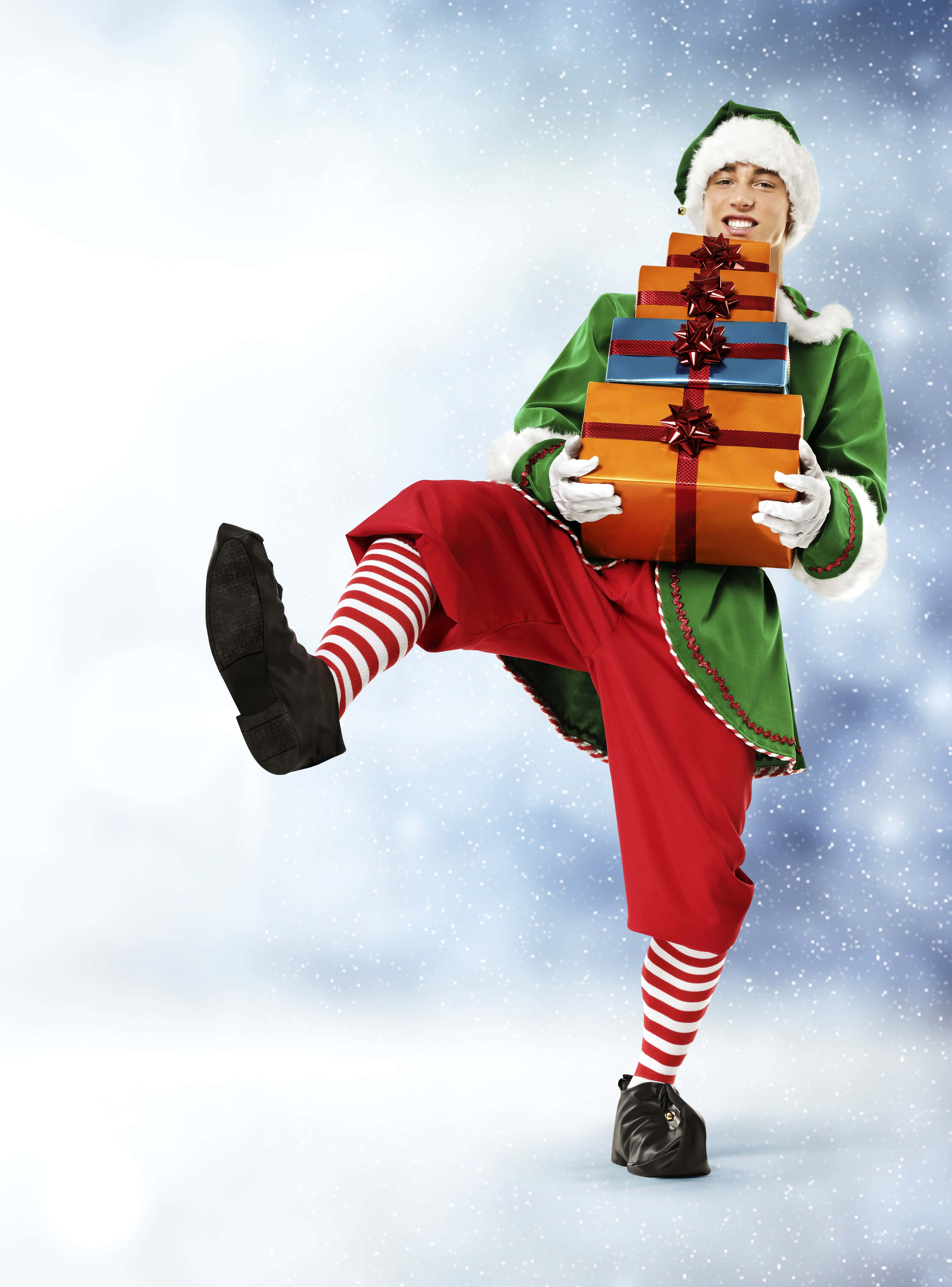 Christmas Elf 桌布 壁紙 — 一個快樂的精靈穿著節日服裝，在雪景背景下充滿活力地踢腿，同時平衡著一堆色彩繽紛的禮物盒。
