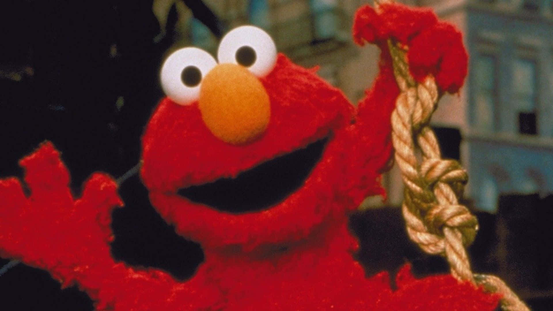 Elmo 桌布 壁紙 — 一個快樂的紅色毛絨公仔，有大大的白色眼睛和橙色鼻子，一邊揮手一邊拿著一條粗繩結。