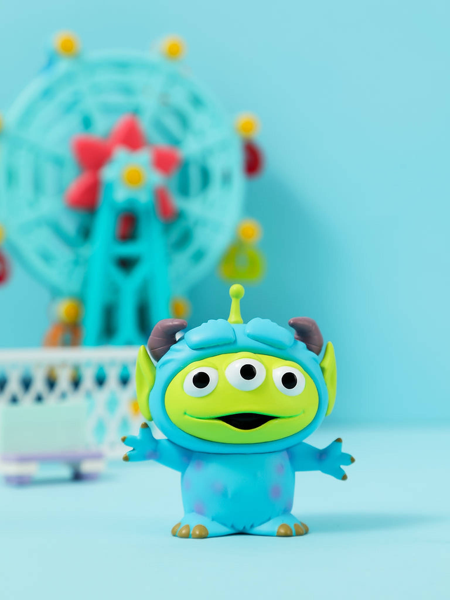 Toy Story Alien 桌布 壁紙 — 一個快樂的藍色三眼外星人玩具站在模糊的藍綠色摩天輪前，背景是柔和的藍色。