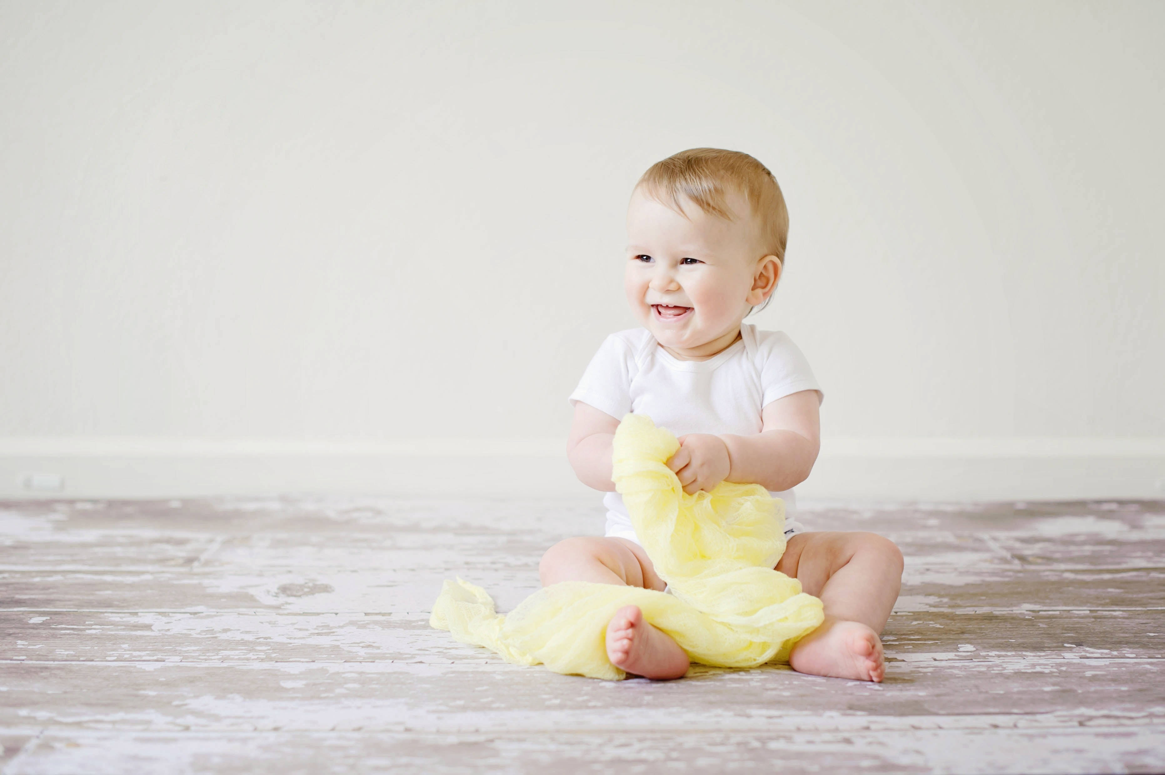 Baby Boy 桌布 壁紙 — 一個快樂的金髮嬰兒坐在風化的木地板上，笑著抓住一塊黃色的布。