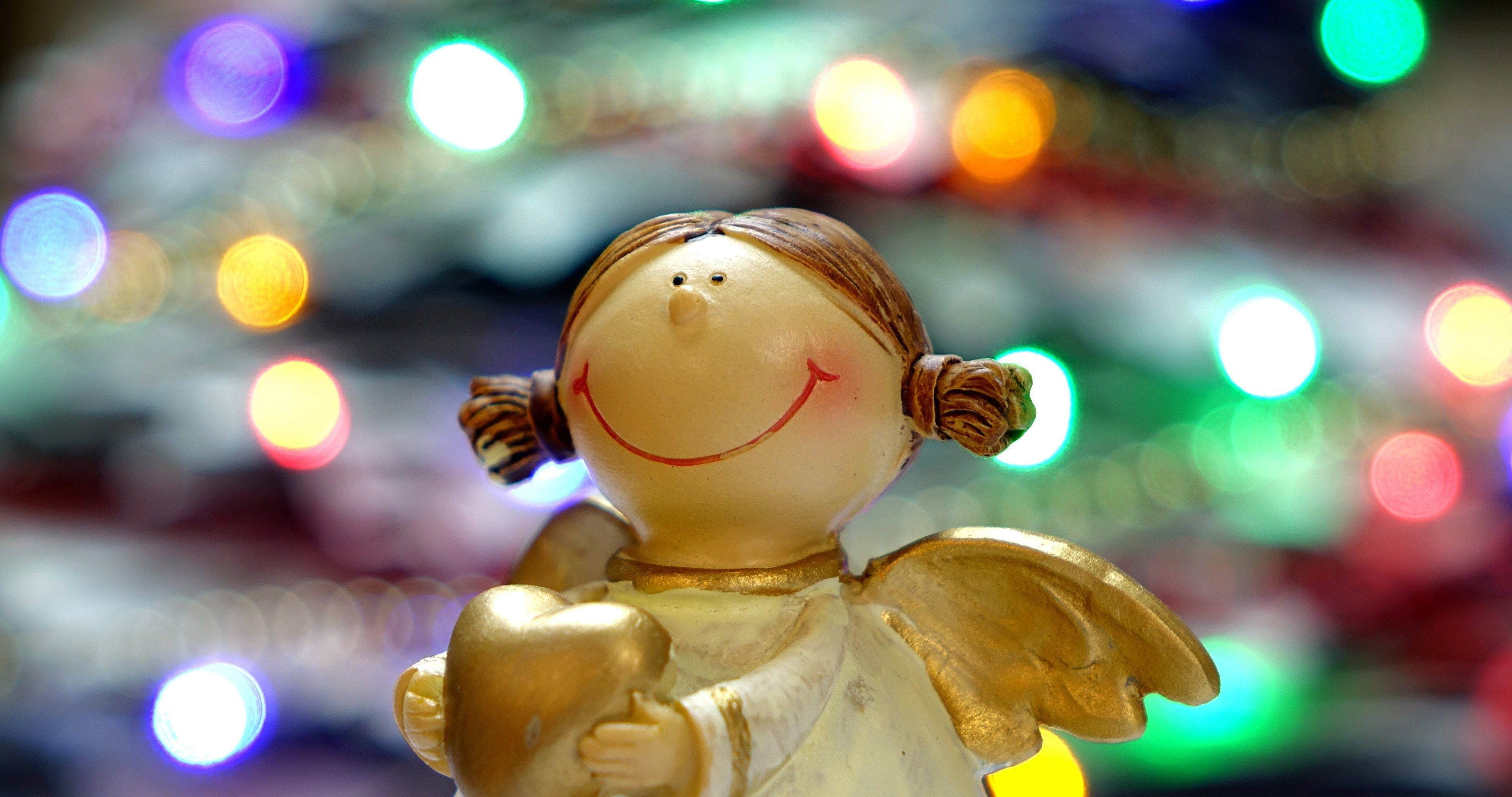 angels 桌布 壁紙 — 一個快樂的陶瓷天使雕像，金色翅膀，在節日燈光的鮮豔散景中手持一顆心。