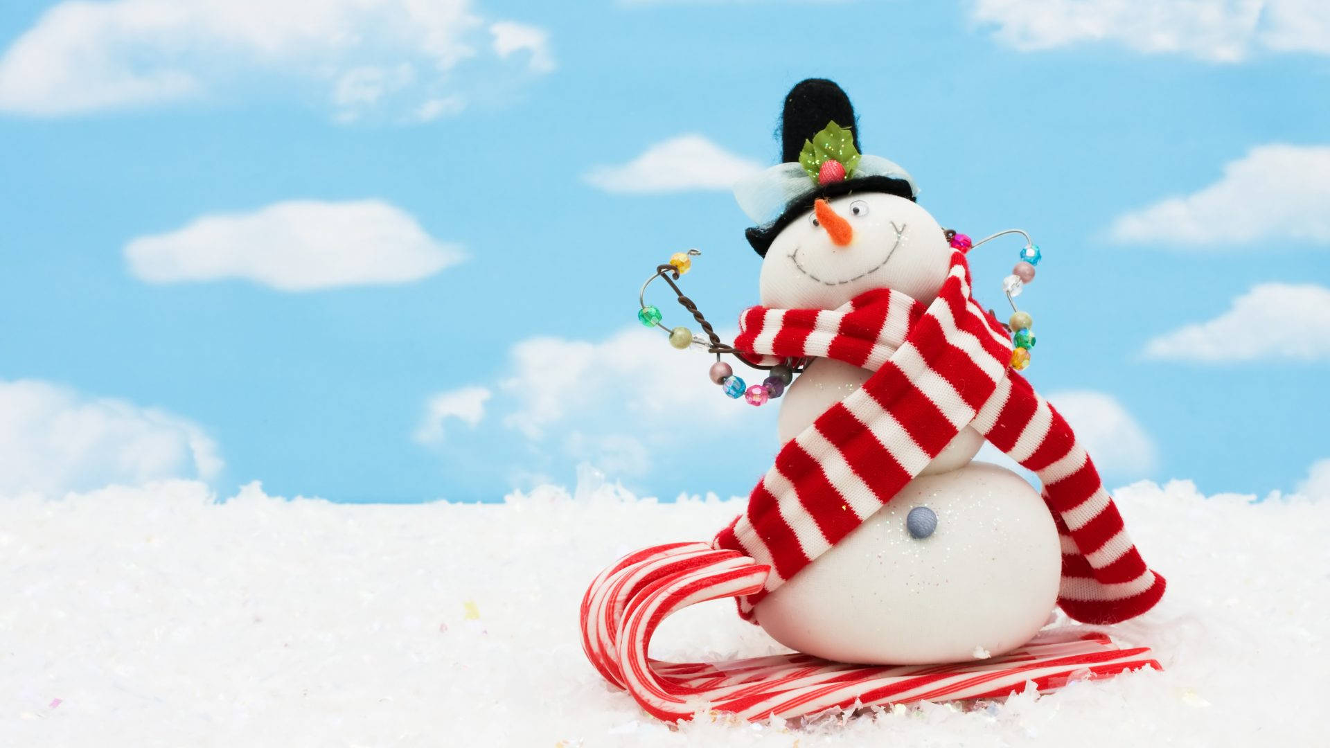 Cute Snow 桌布 壁紙 — 一個快樂的雪人，戴著紅白條紋圍巾和拐杖糖雪橇，坐在蓬鬆的雪地上，天空湛藍，雲朵稀疏。