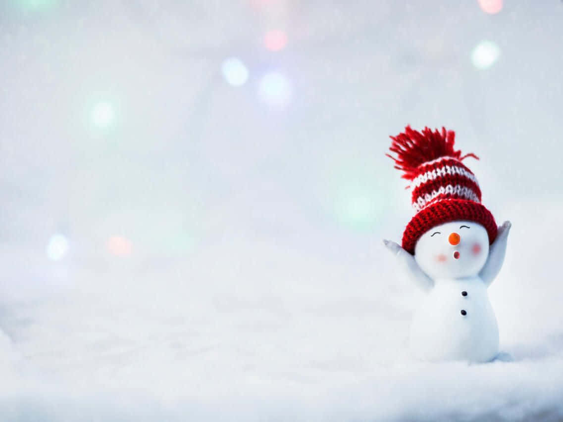 Teams Christmas 桌布 壁紙 — 一個快樂的雪人戴著紅白相間的針織帽，帽頂有絨球，站在雪地上，背景是柔和發光的燈光。