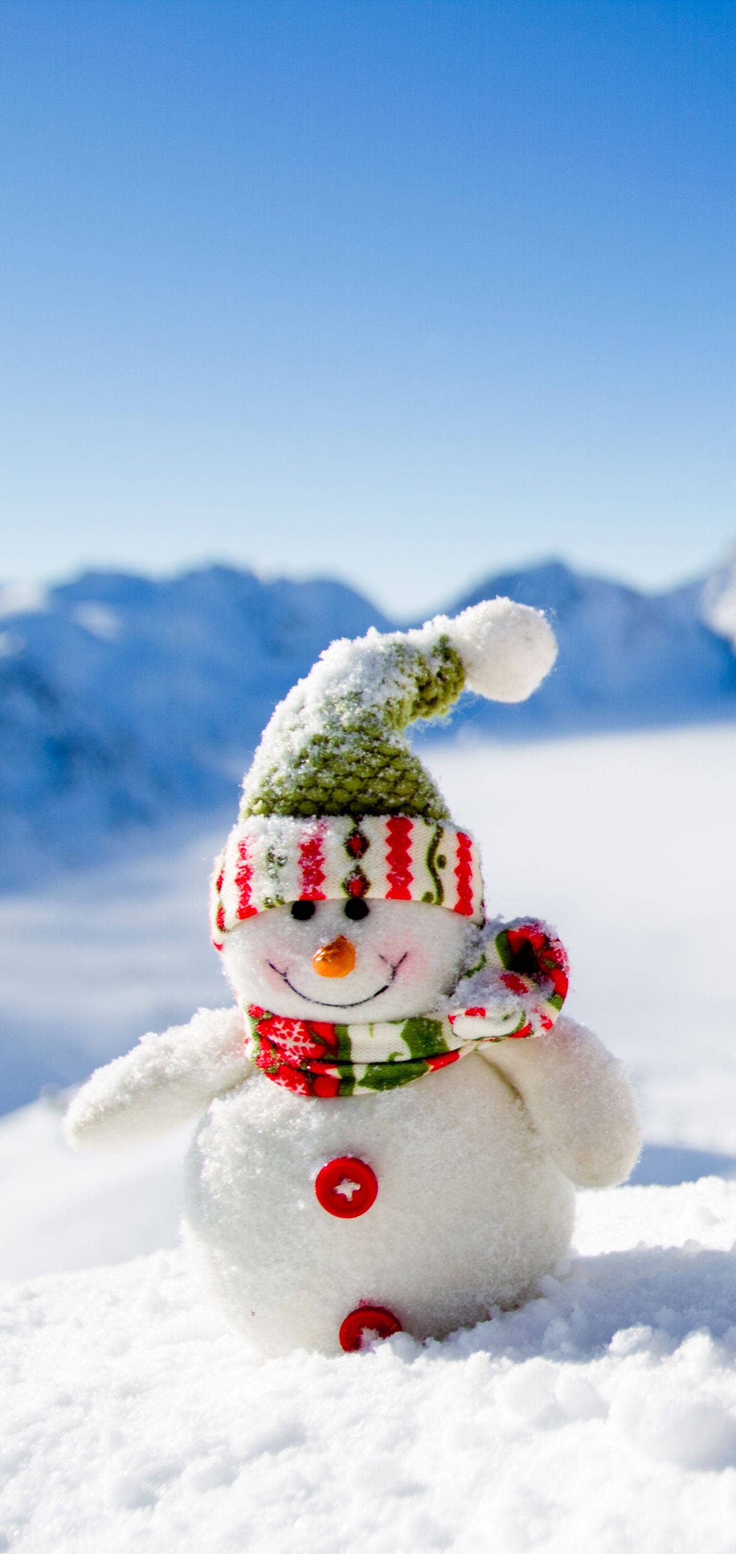 冬季 iPhone 壁紙 桌布 壁紙 — 一個快樂的雪人戴著綠色針織帽和紅色圍巾，站在白雪覆蓋的田野上，遠處有山脈，天空清澈湛藍。