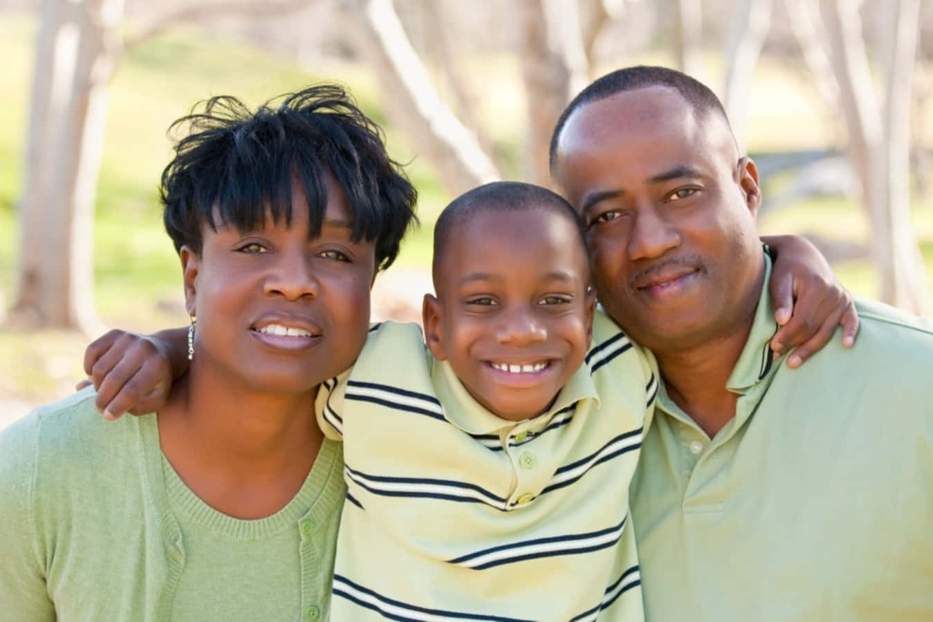 Black Family 桌布 壁紙 — 一個快樂的非裔美國三人家庭在明亮的陽光下，背景有樹木，在戶外溫暖地微笑。