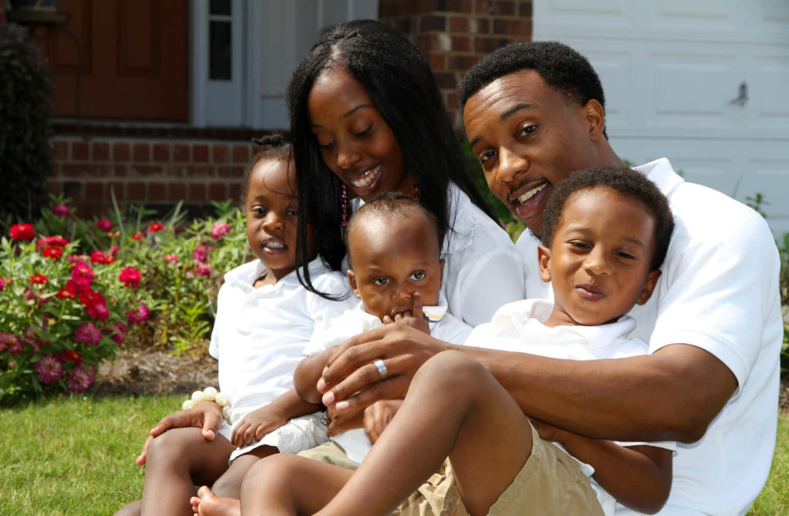 Black Family 桌布 壁紙 — 一個快樂的非裔美國五人家庭一起坐在他們屋前的草坪上，在紅花點綴的磚房前溫暖地微笑。