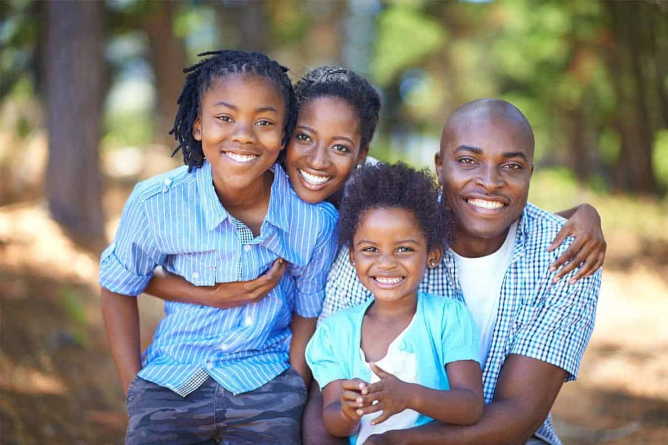 Black Family 桌布 壁紙 — 一個快樂的非裔美國四人家庭在陽光明媚的森林空地中溫暖地微笑，斑駁的光線穿過樹木。
