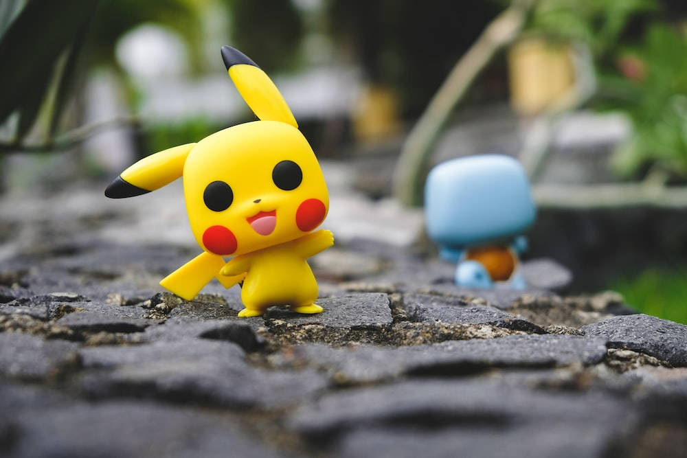 Squirtle 桌布 壁紙 — 一個快樂的黃色皮卡丘公仔站在鵝卵石小路上，背景模糊中有一個藍色傑尼龜公仔。