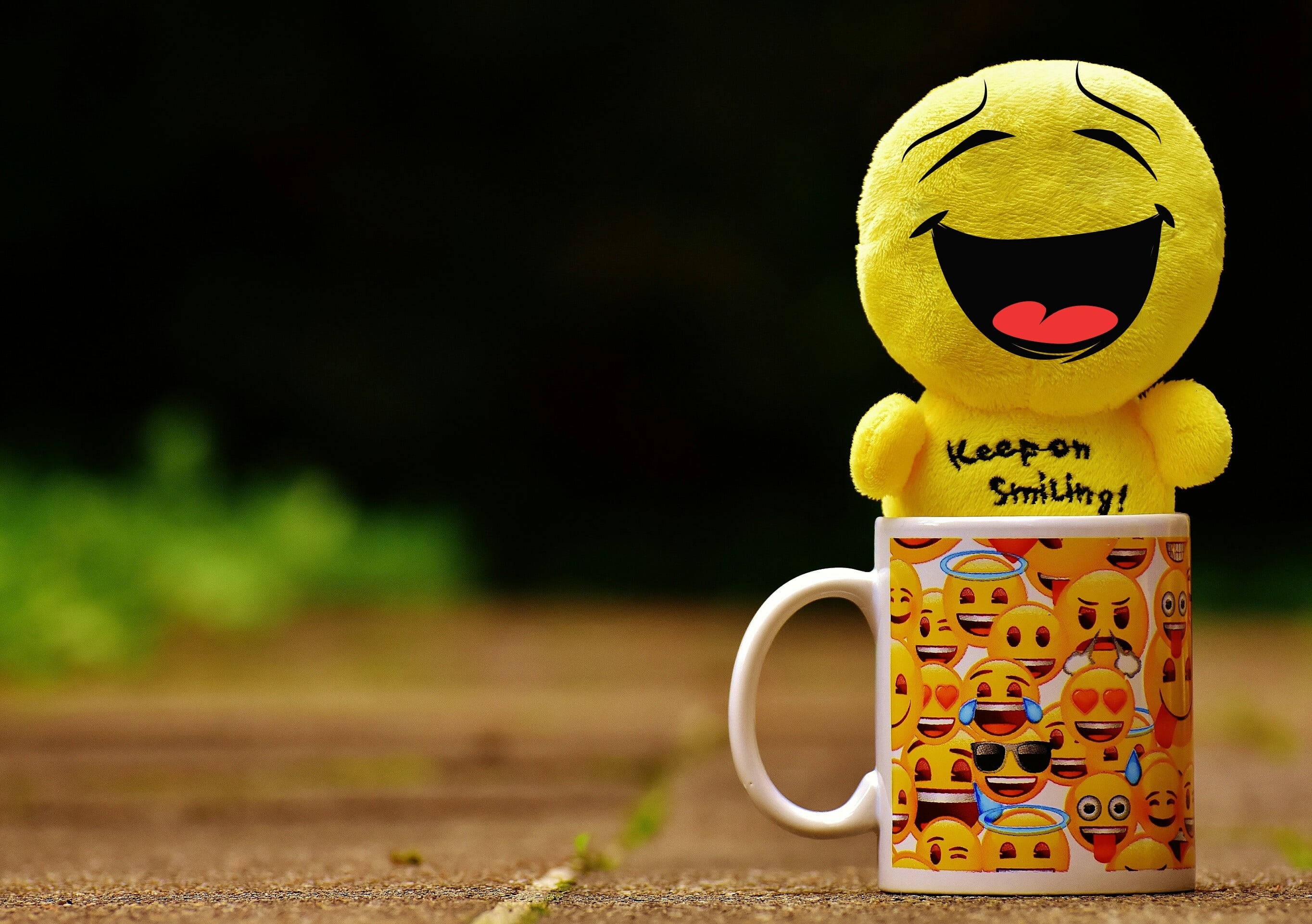laughing emoji 桌布 壁紙 — 一個快樂的黃色笑臉表情公仔放在一個佈滿彩色表情圖案的杯子上。