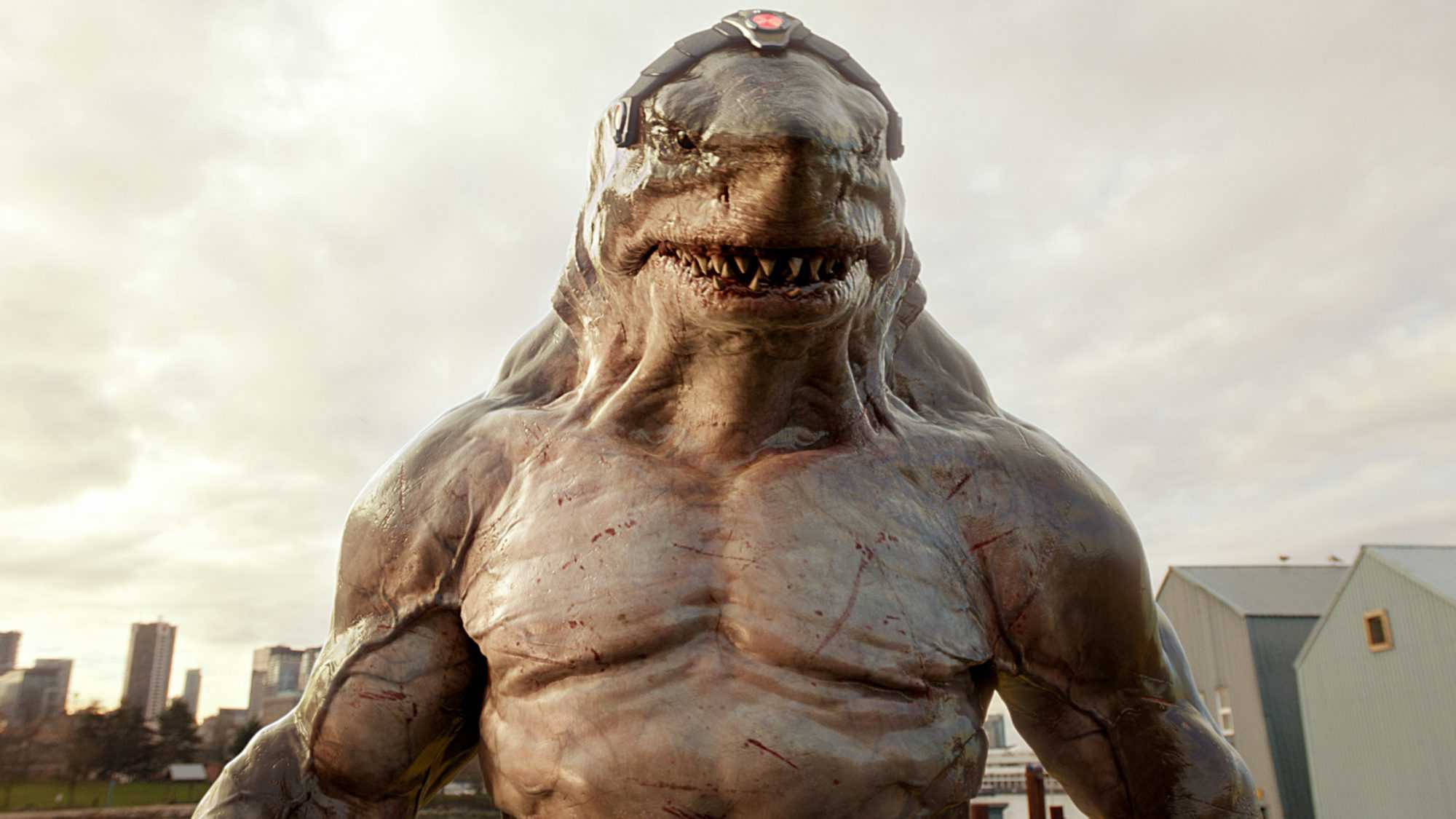 King Shark 桌布 壁紙 — 一個怪獸般的鯊魚頭人形生物，皮膚血淋淋，戴著頭盔，在陰雲密佈的城市天際線前威嚇地站立。