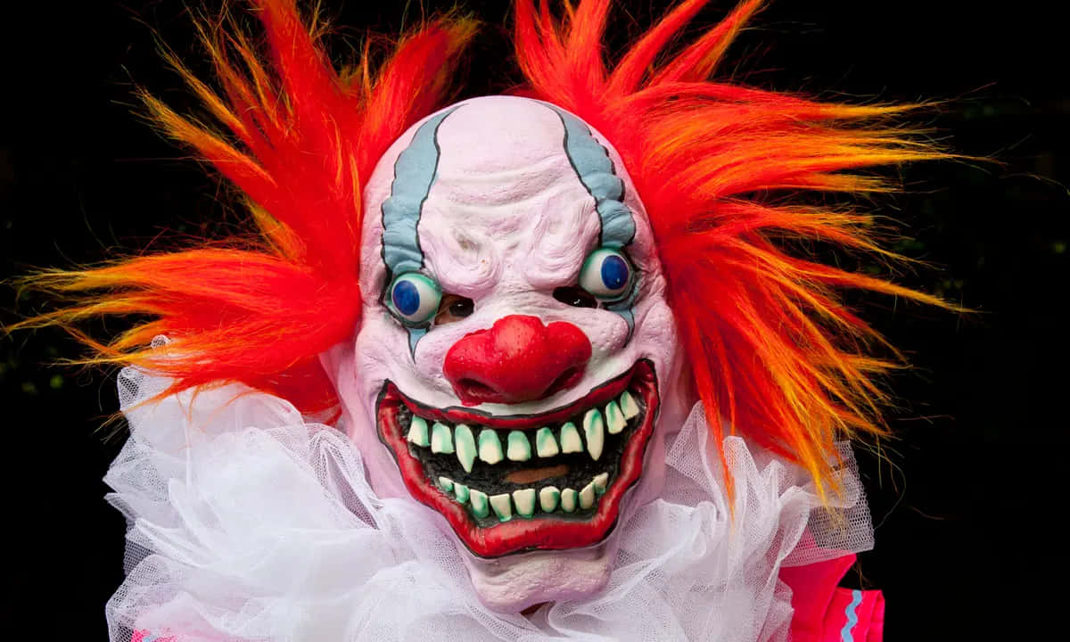 Creepy Clown 桌布 壁紙 — 一個怪誕的小丑面具，有著狂野的橙色頭髮和鋒利的牙齒，在黑色背景上強烈地凝視著。