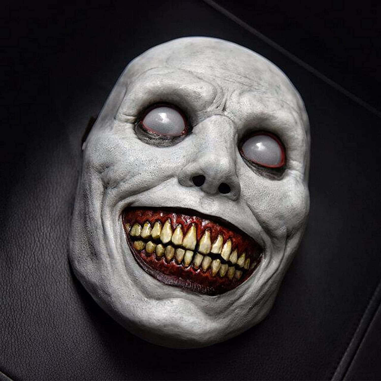 Creepy Halloween 桌布 壁紙 — 一個怪誕蒼白的面具，帶有凸出的紅邊眼睛和露出泛黃牙齒的咧嘴笑容，放置在黑色皮革表面上。