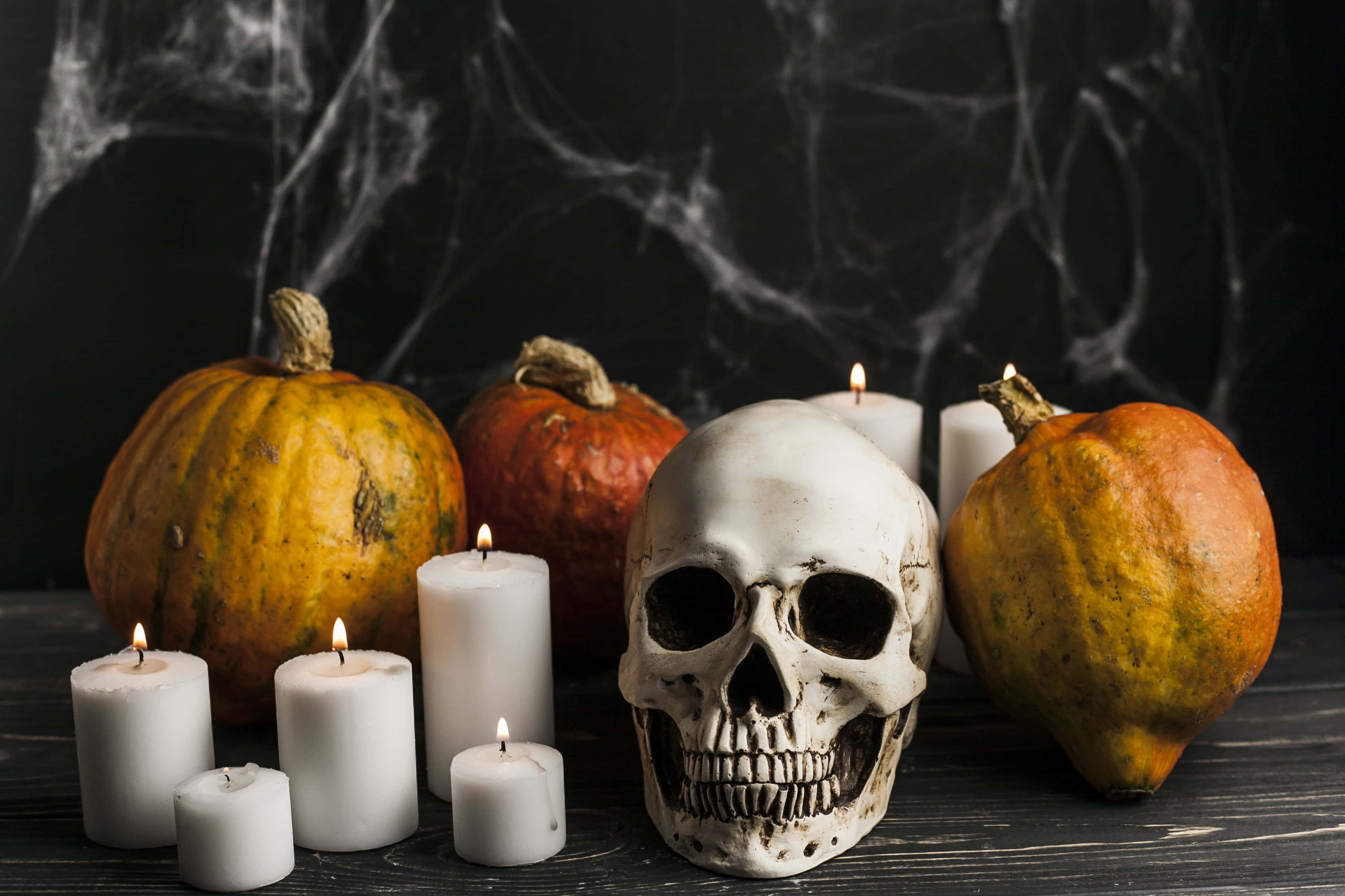 Halloween Props 桌布 壁紙 — 一個恐怖的萬聖節靜物畫，有一個人類頭骨被南瓜和閃爍的白色蠟燭包圍，背景是黑暗的蜘蛛網。