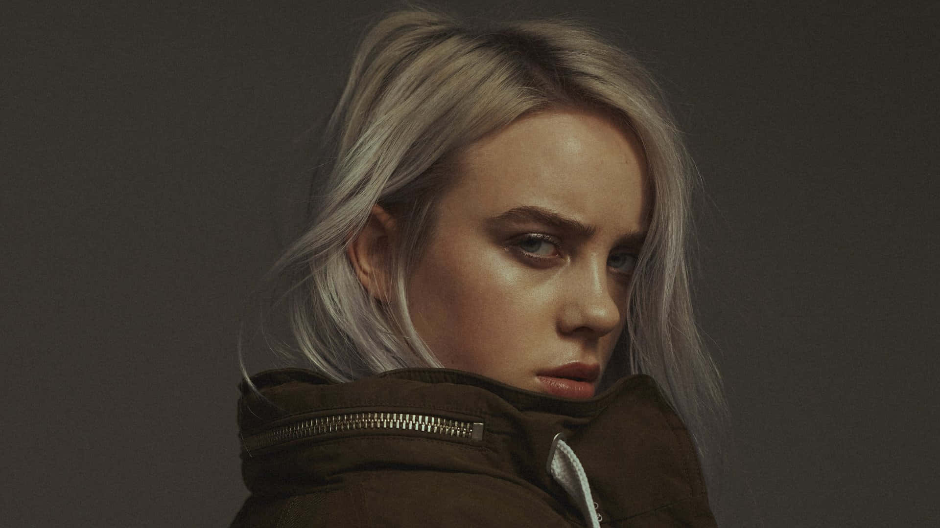 Billie Eilish 2021 桌布 壁紙 — 一個情緒化的肖像，一位金髮女子穿著深色外套，在灰色背景下凝視著。