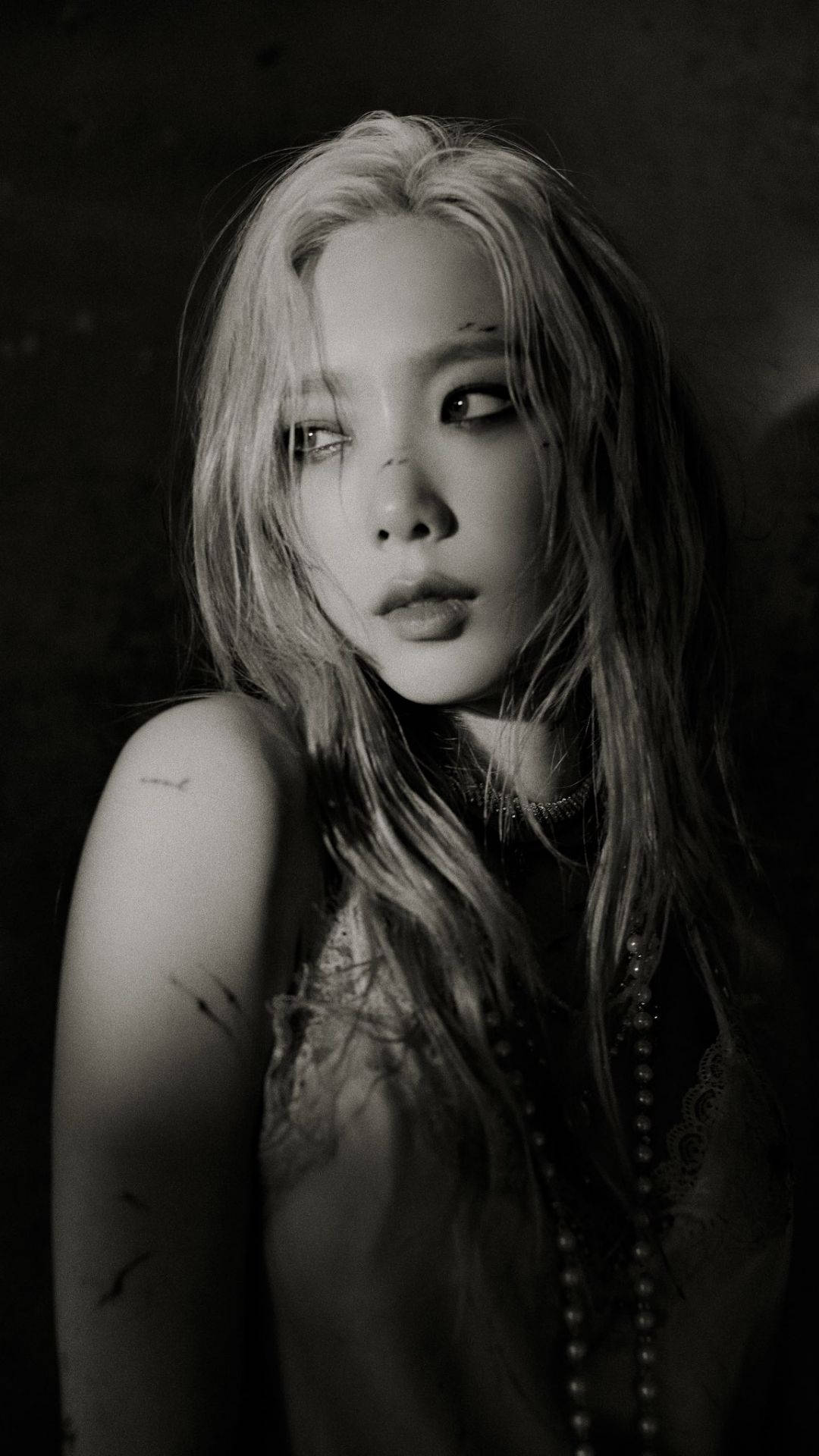 Taeyeon 桌布 壁紙 — 一個情緒化的黑白肖像，描繪一位長髮且有刺青的女性，在深色紋理背景前沉思地凝視。