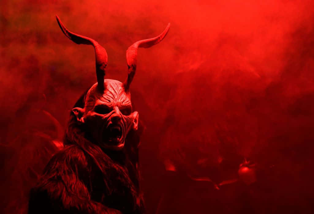 Krampus 桌布 壁紙 — 一個惡魔般的身影，長著彎曲的角和皺起的眉頭，在戲劇性燈光下，於旋轉的紅色煙霧中咆哮。