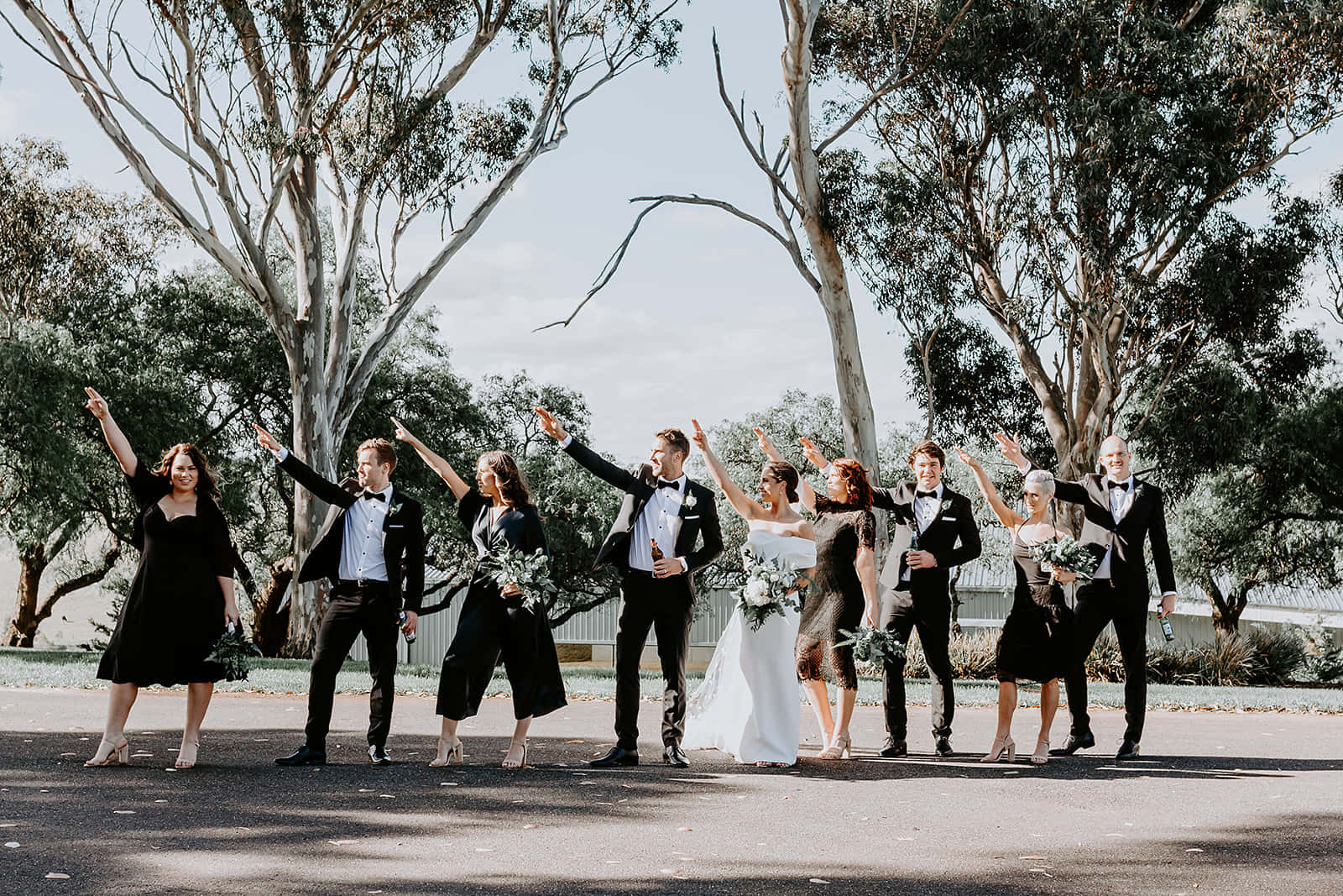 Bridal 桌布 壁紙 — 一個愉快的婚禮派對穿著正式服裝，在晴朗的日子裡，在高大的桉樹下舉起雙臂擺姿勢。