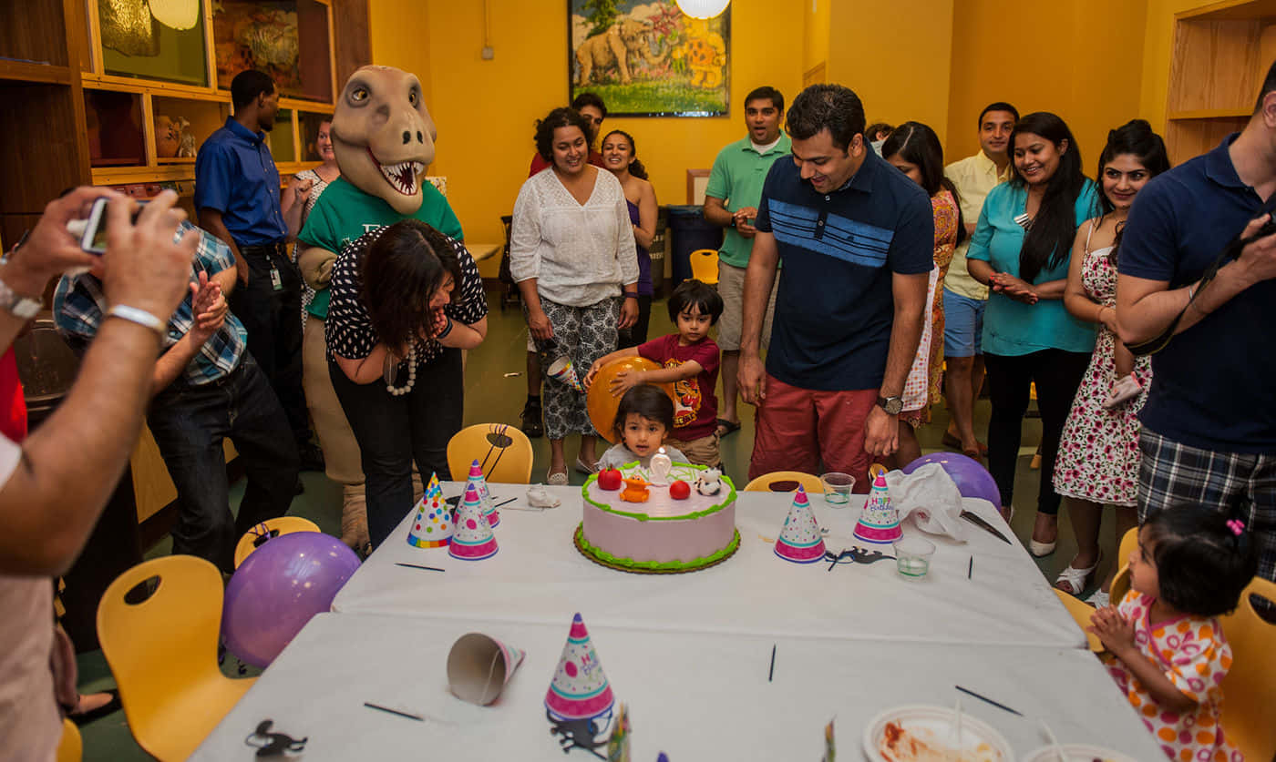 Birthday Party 桌布 壁紙 — 一個愉快的生日派對，孩子在家人和穿著恐龍服裝的人圍繞下吹熄蛋糕上的蠟燭。