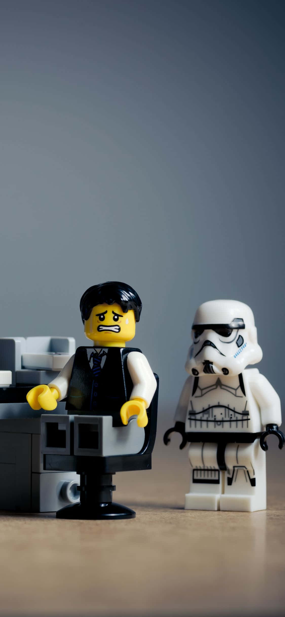 背景 桌布 壁紙 — 一個憤怒表情的LEGO商人坐在椅子上，旁邊有一個LEGO風暴兵，背景為灰色。