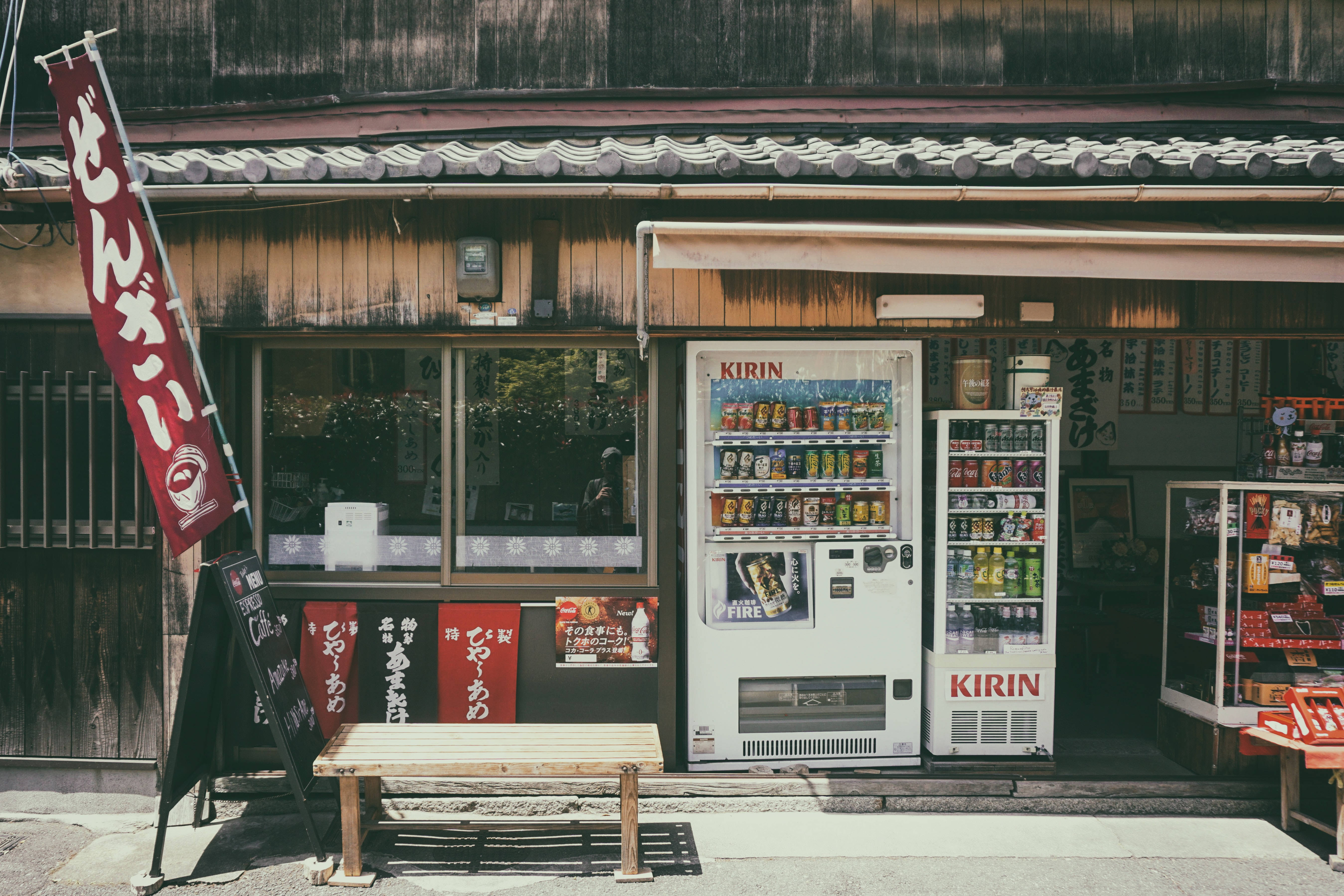 日本 桌布 壁紙 — 一個懷舊日本街角，在溫暖陽光下，木長椅和傳統店鋪旁邊有兩台 Kirin 自動販賣機。