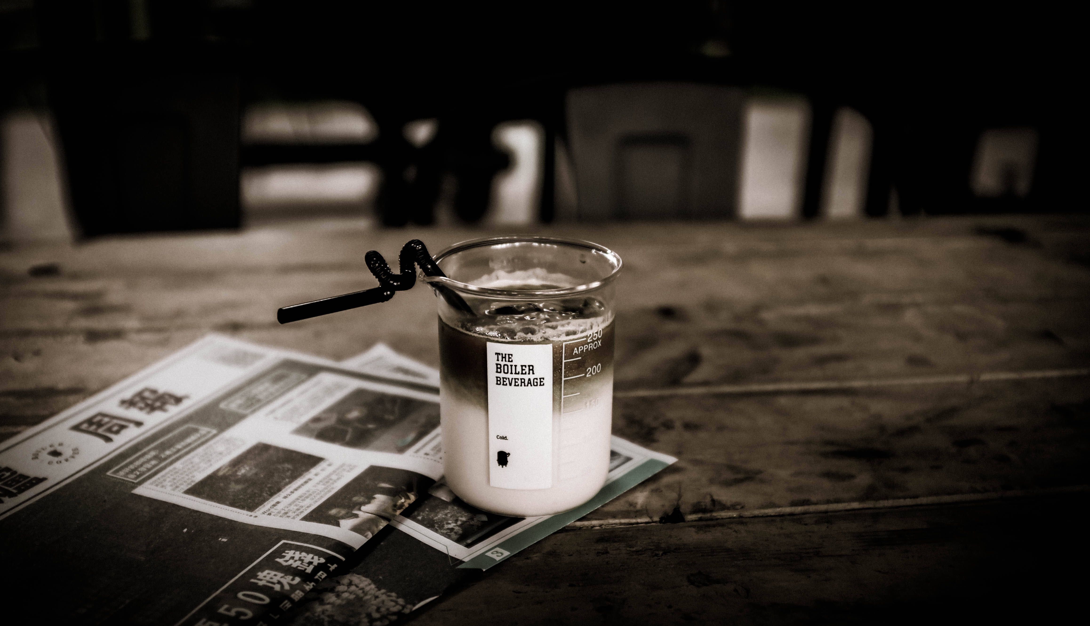 Beaker 桌布 壁紙 — 一個懷舊色調的燒杯，標有「THE BOILER BEVERAGE」，帶有黑色吸管，放在一張舊木桌上的報紙上。