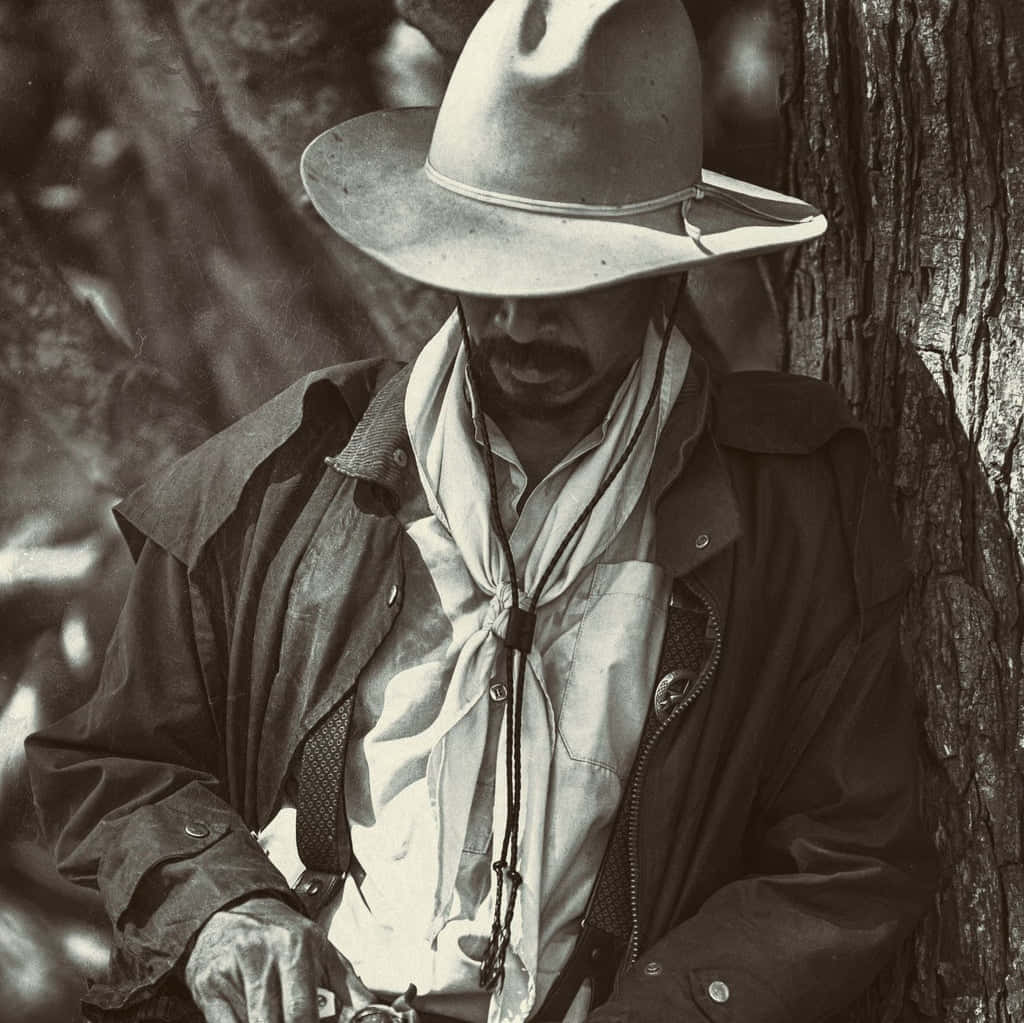 Cowboy Hat 桌布 壁紙 — 一個懷舊色調的牛仔戴著寬邊帽，靠在一棵樹幹上，背景是崎嶇的戶外環境。