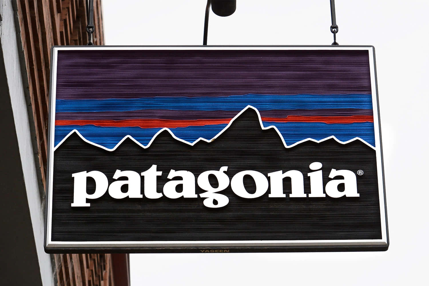 Patagonia 桌布 壁紙 — 一個懸掛的Patagonia商店招牌，特色是風格化的山脈，帶有紫色、藍色和紅色條紋，背景為黑色。