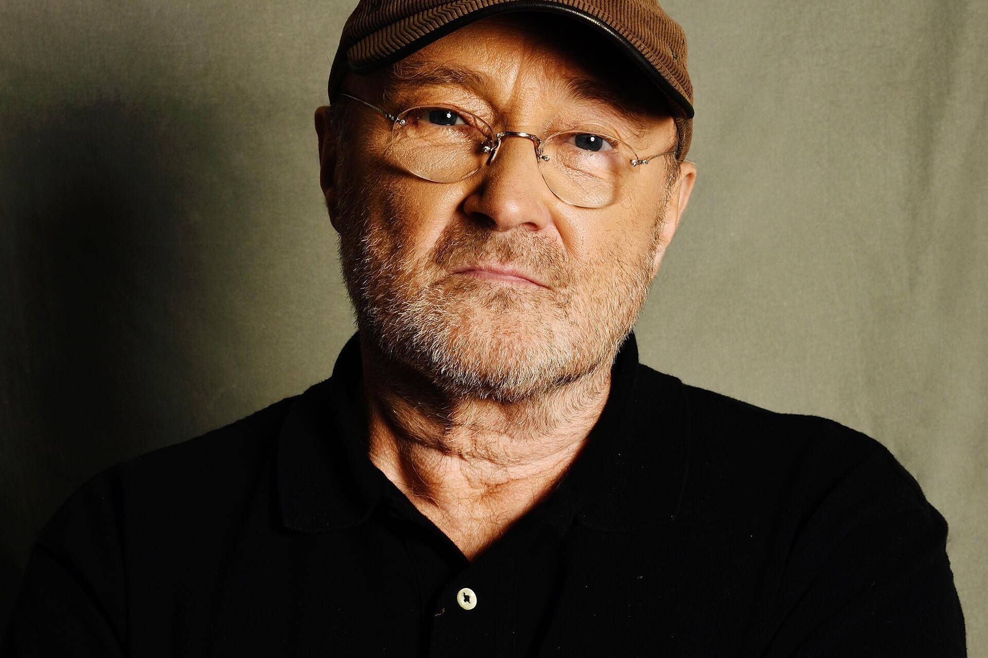 phil collins 桌布 壁紙 — 一個成熟男人戴著眼鏡和帽子的特寫肖像，背景為柔和的灰色。