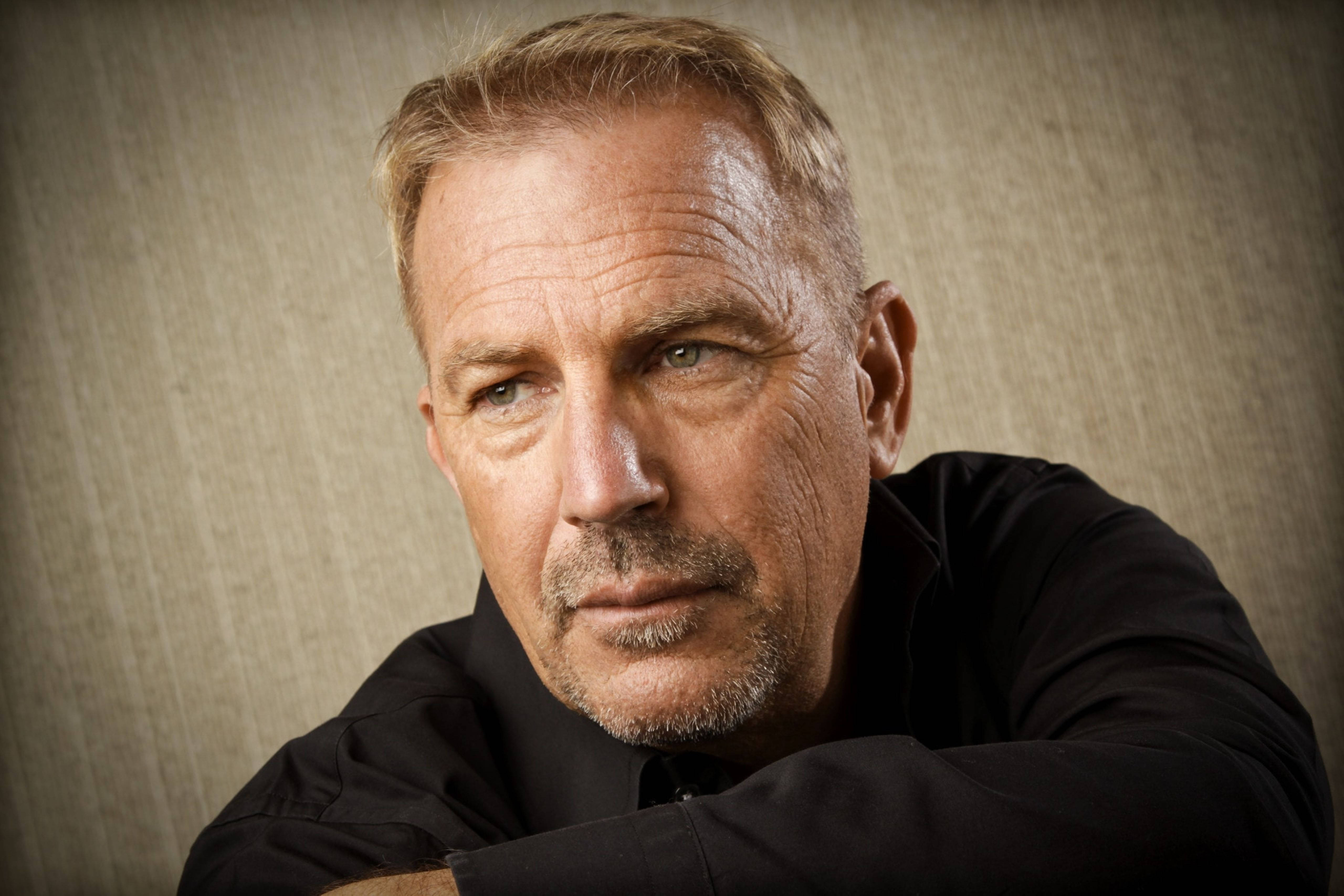 Kevin Costner 桌布 壁紙 — 一個成熟男人的特寫肖像，灰白頭髮和鬍鬚，穿著黑色襯衫，背景是帶紋理的米色。