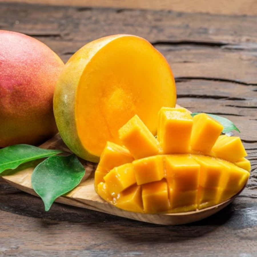 Mango 桌布 壁紙 — 一個成熟的芒果切成多汁的方塊，旁邊放著整個水果和綠葉，放在質樸的木板上。