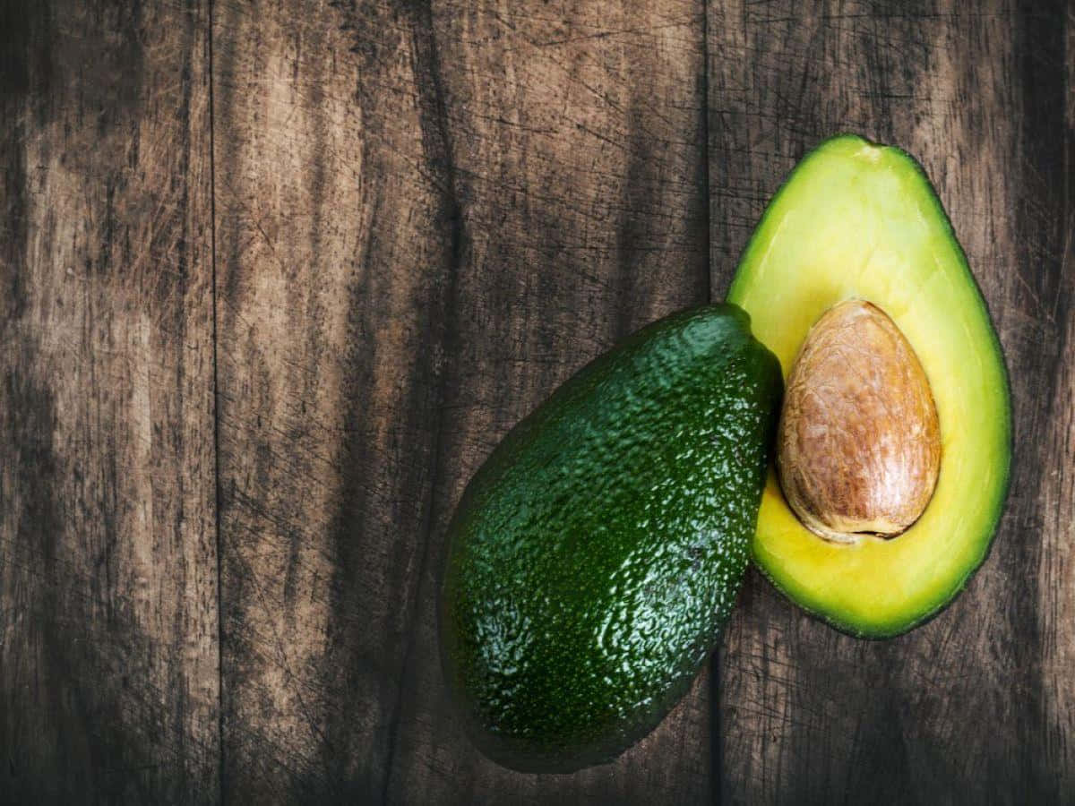 Avocado 桌布 壁紙 — 一個成熟的酪梨切半，露出其奶油狀的綠色果肉和大的棕色果核，放在深色質樸的木質表面上。