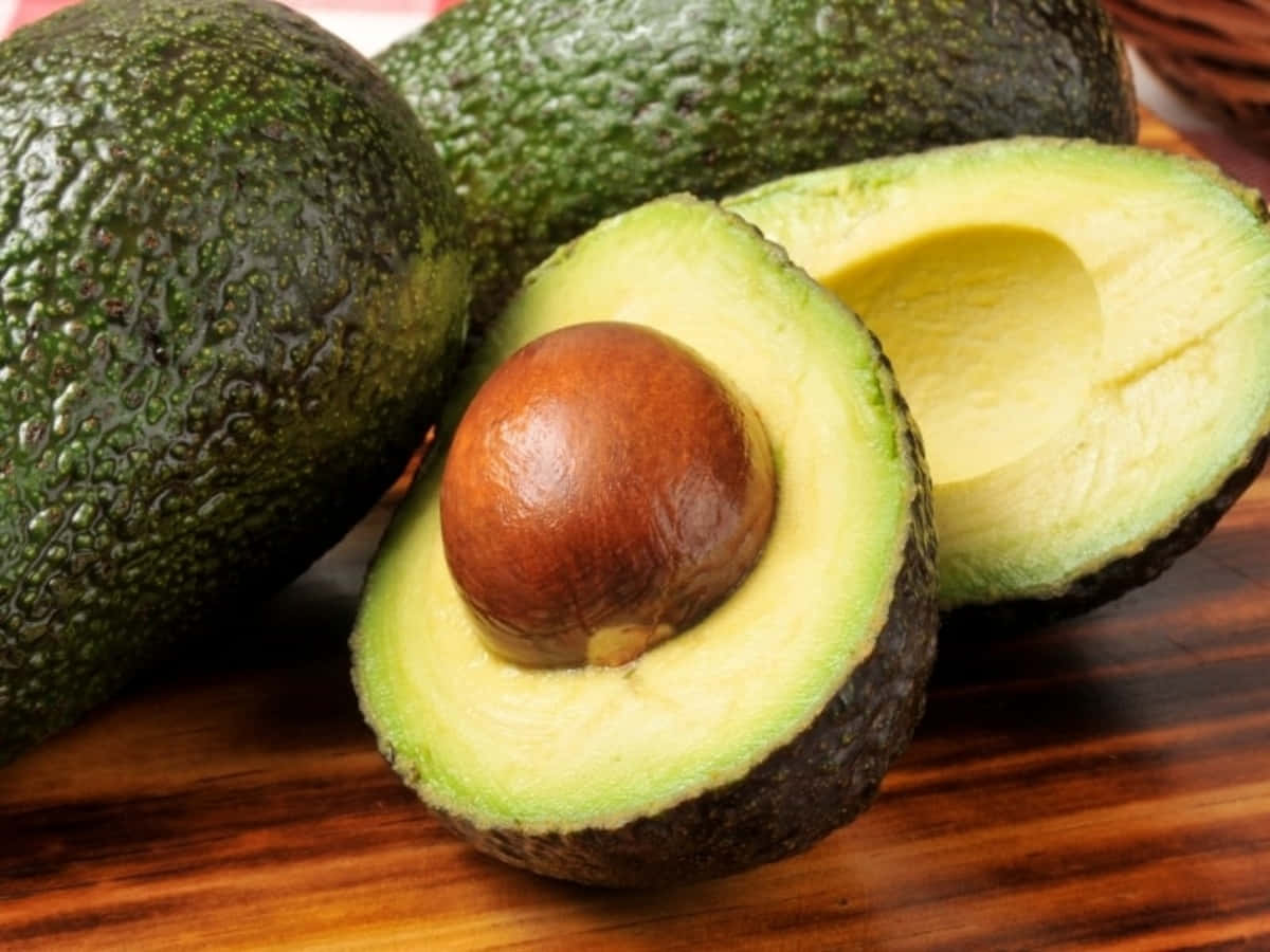 Avocado 桌布 壁紙 — 一個成熟的酪梨切半，露出奶油狀的綠色果肉和一個大的棕色果核，放在木質表面上。