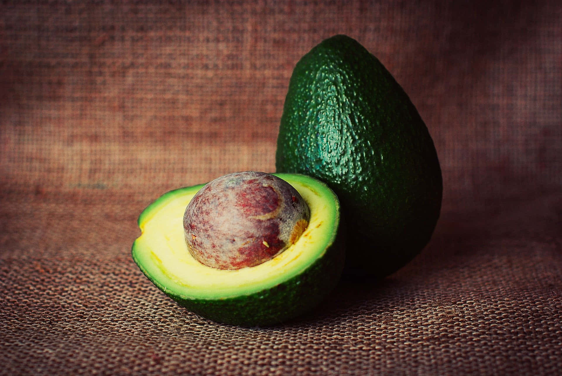 Avocado 桌布 壁紙 — 一個成熟的酪梨切半，露出奶油黃色的果肉和大的棕色種子，放在有紋理的棕色粗麻布表面上。