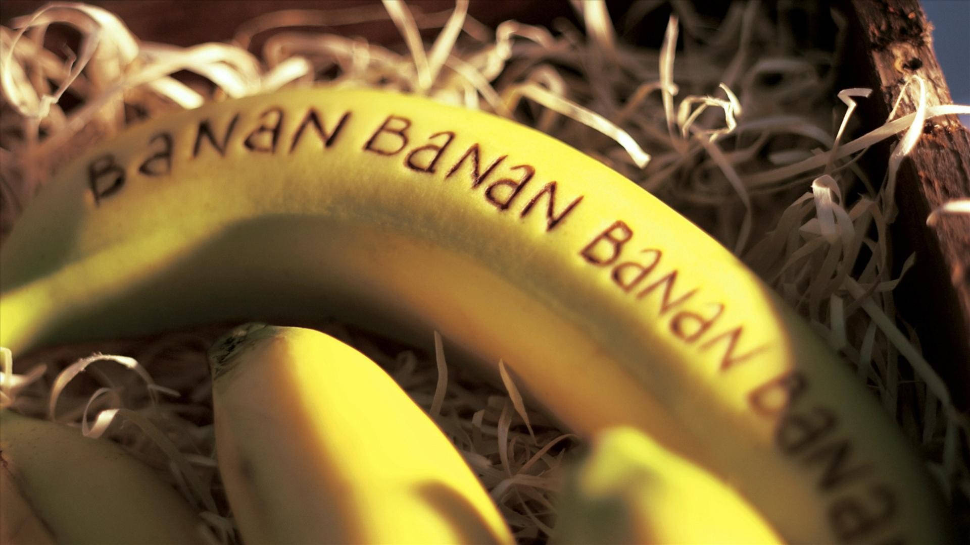 香蕉 桌布 壁紙 — 一個成熟的香蕉，果皮上刻有 'BANAN'，躺在沐浴在溫暖金色光線中的稻草上。