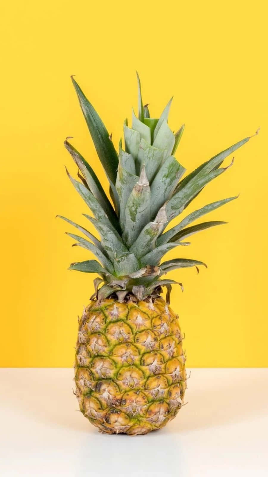 Pineapple 桌布 壁紙 — 一個成熟的鳳梨帶有尖刺的綠色冠部，位於白色表面上，背景為鮮黃色。