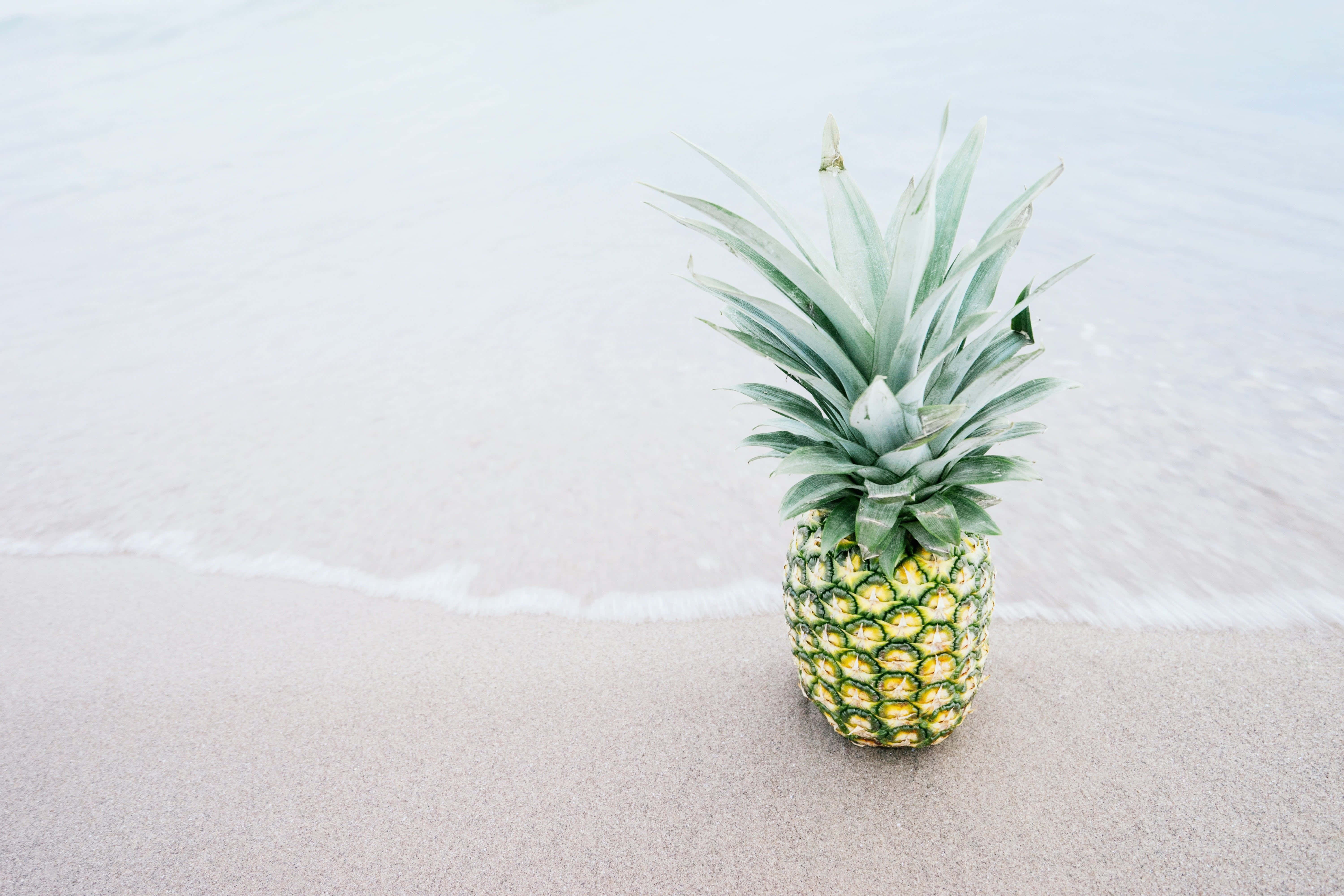 Pineapple 桌布 壁紙 — 一個成熟的鳳梨放在柔軟的沙上，溫柔的海浪在明亮的日光下拍打著海岸。