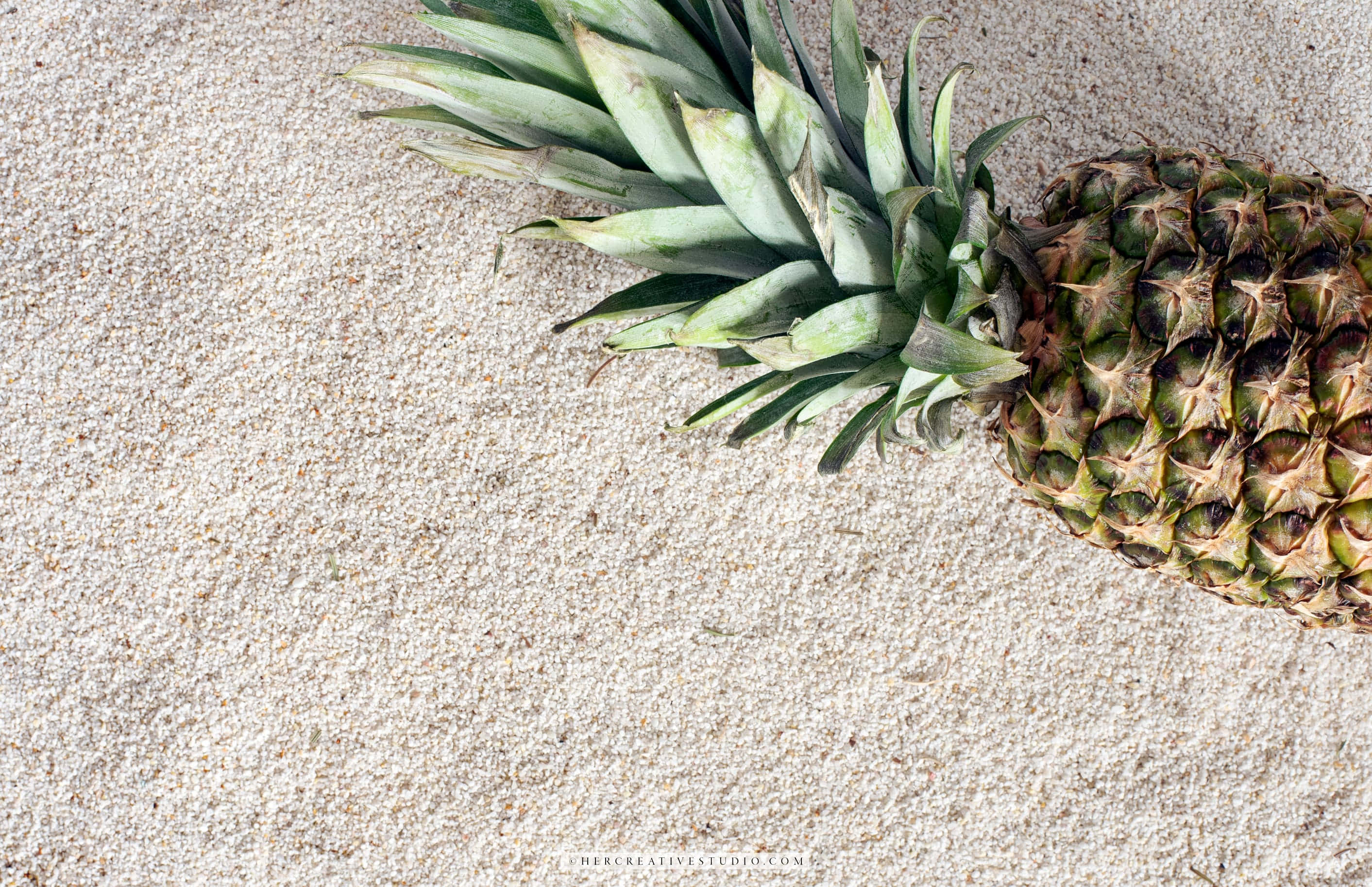 Pineapple Desktop 桌布 壁紙 — 一個成熟的鳳梨放在淺米色沙子上，其帶刺的綠色冠部朝上。