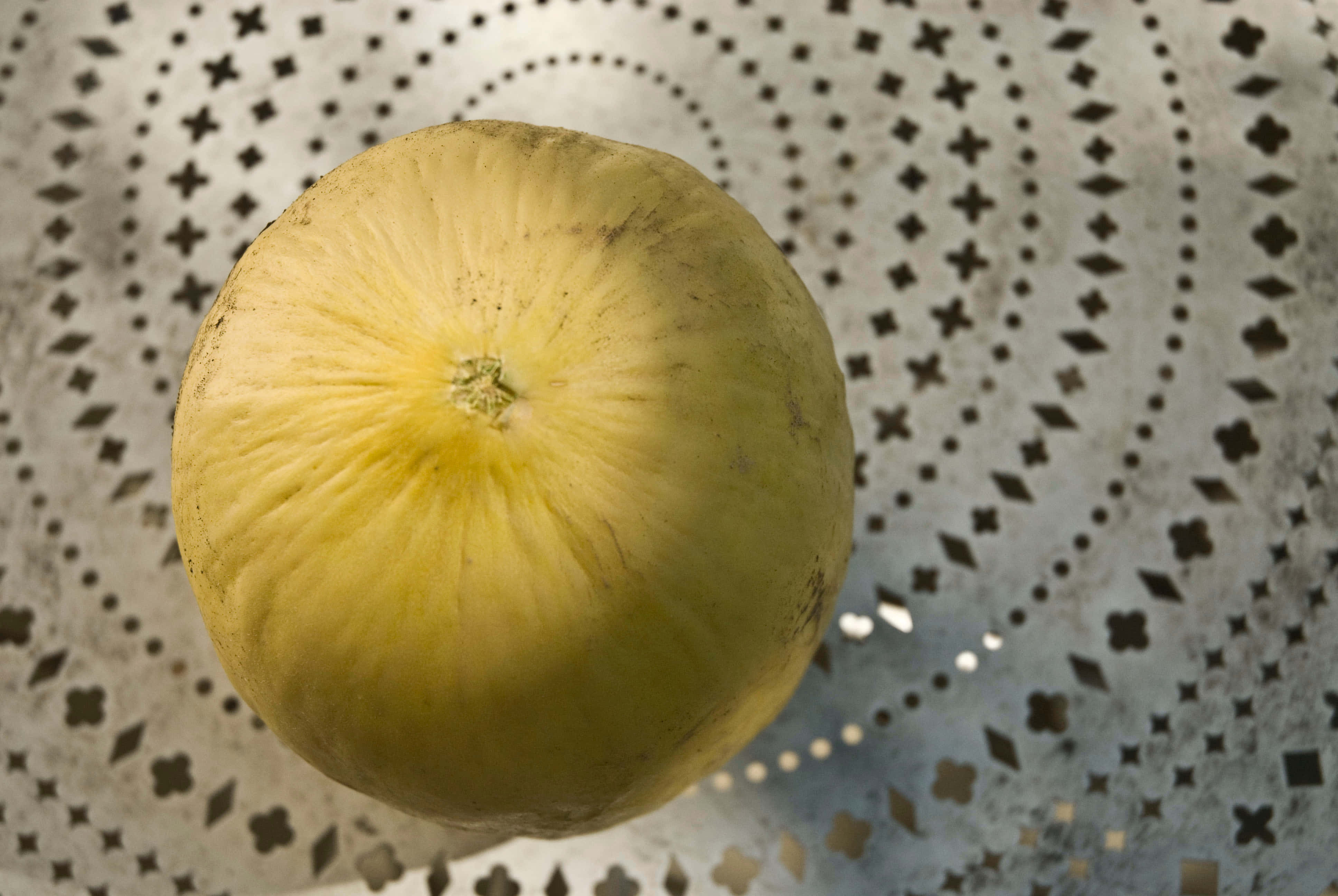 Food Photography 桌布 壁紙 — 一個成熟的黃色哈密瓜放在一個帶有星形孔洞的圖案金屬濾鍋上。