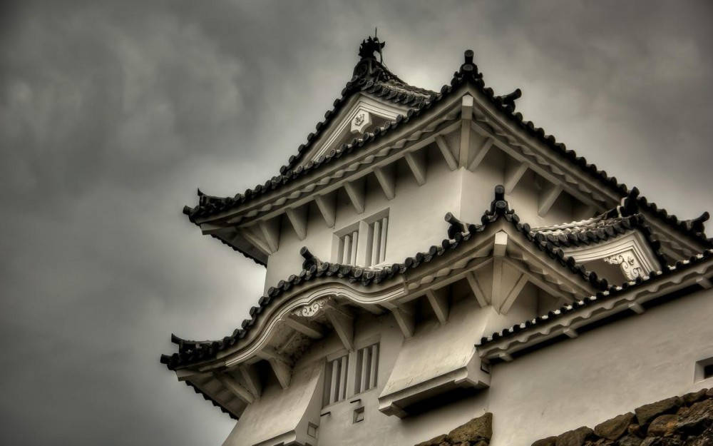 Himeji Castle 桌布 壁紙 — 一個戲劇性的低角度拍攝，傳統日本城堡塔樓，白色牆壁和深色瓦片屋頂，背景是暴風雨的灰色天空。