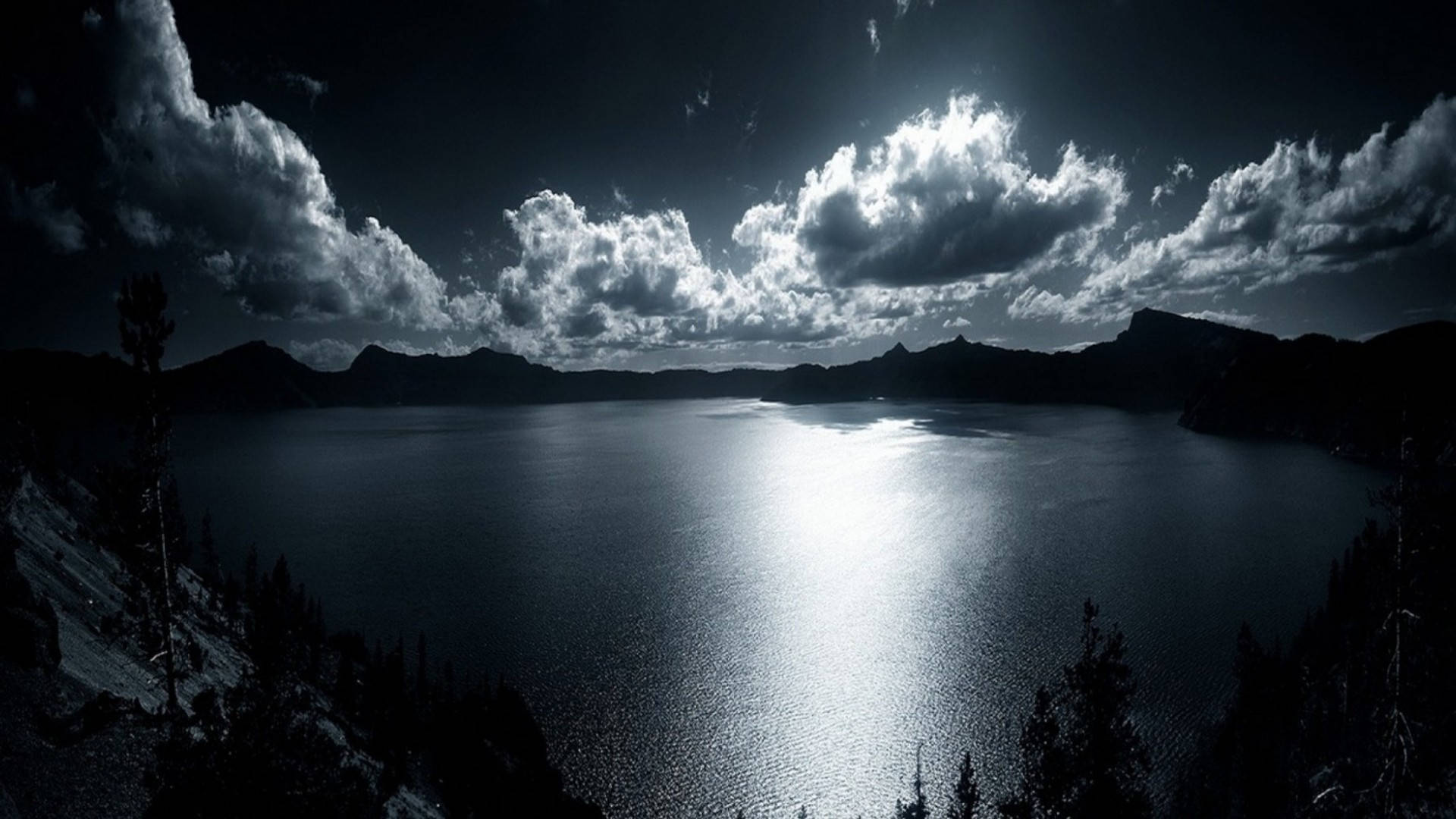 Moonlight 桌布 壁紙 — 一個戲劇性的單色視圖，展示了一個廣闊的火山口湖，在黑暗的天空下反射出明亮的雲彩，並以剪影山脈和樹木為框架。