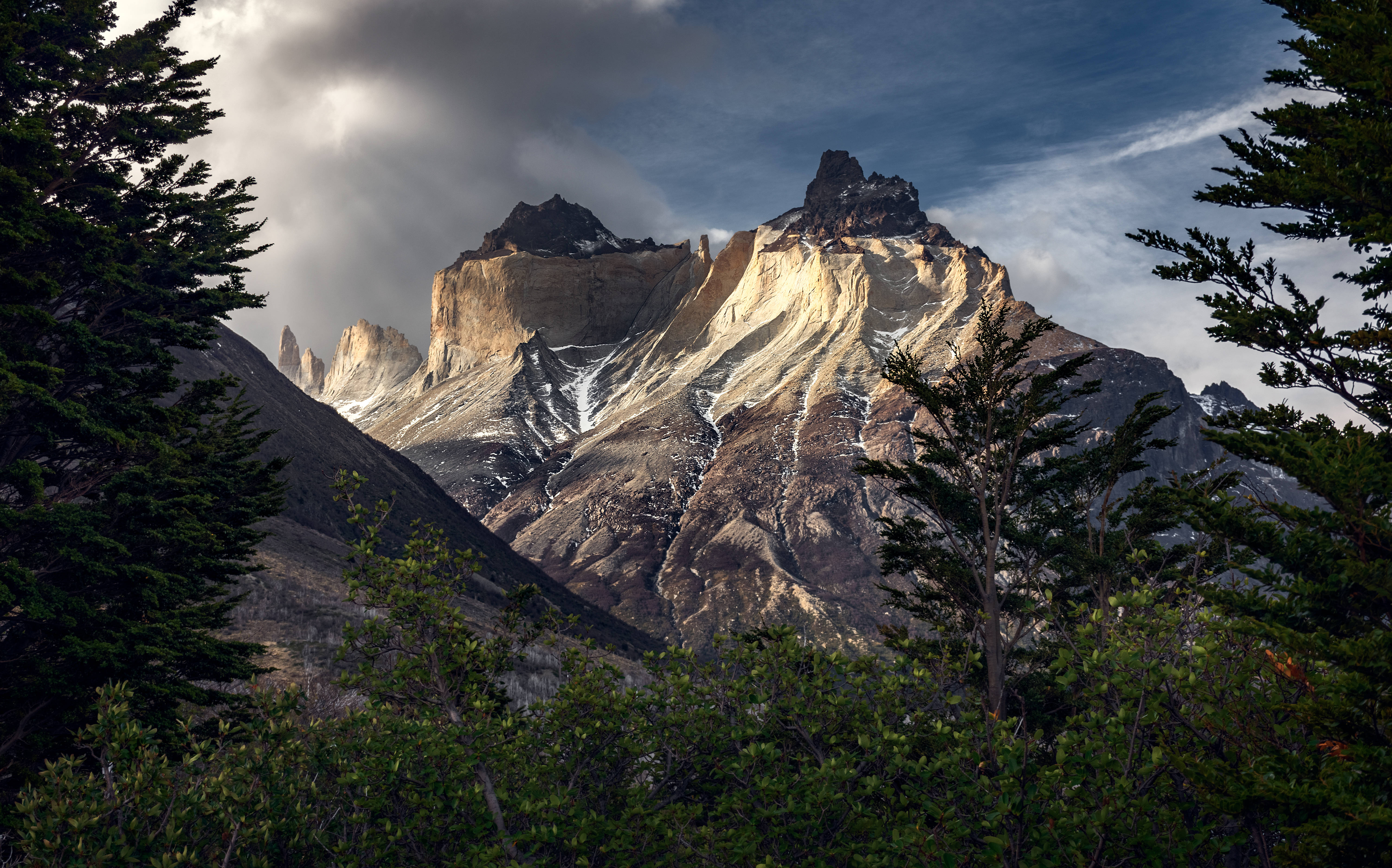 Chile 桌布 壁紙 — 一個戲劇性的山峰，帶有層狀岩石面和雪斑，穿過陰雲密佈的天空，被深色常綠樹環繞。