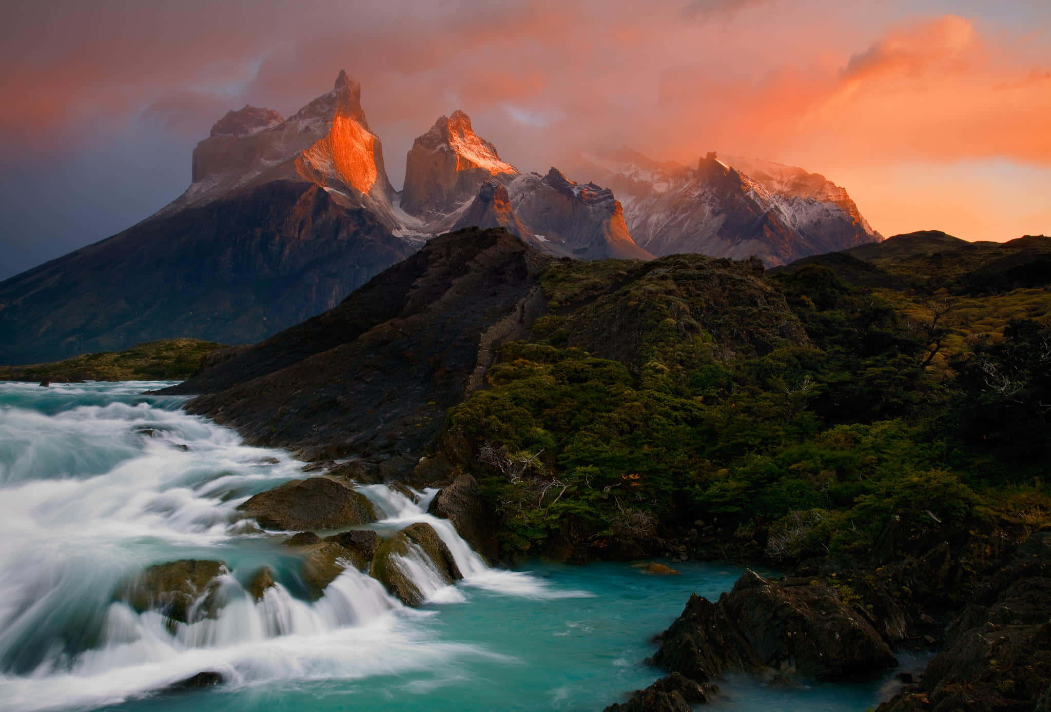 Patagonia 桌布 壁紙 — 一個戲劇性的山峰沐浴在熾熱的日落光芒中，聳立在穿過茂密綠色森林的湍急碧綠河流之上。
