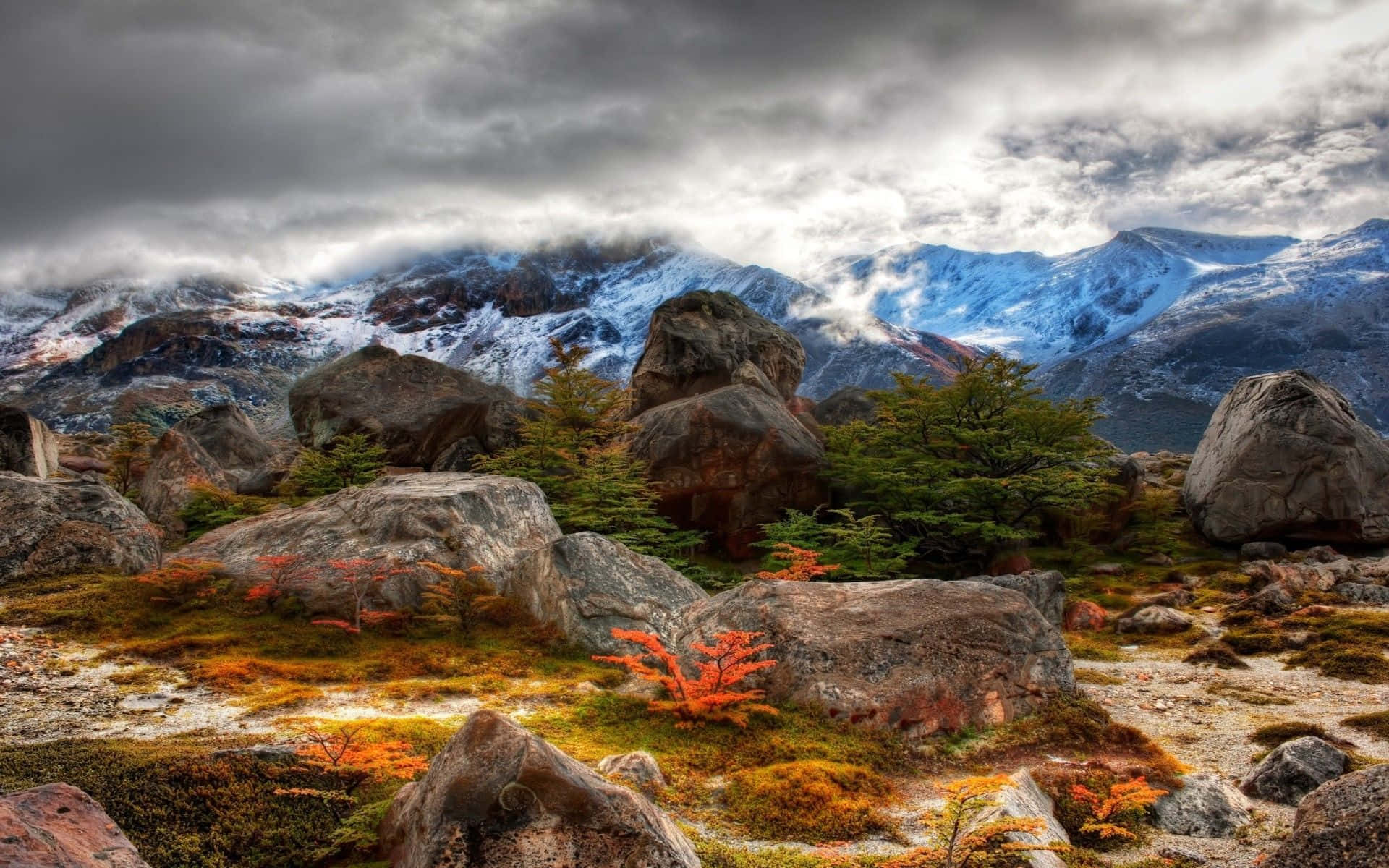 風景 桌布 壁紙 — 一個戲劇性的山景，有白雪覆蓋的山峰、巨大的岩石和陰天天空下充滿活力的橙色樹葉。