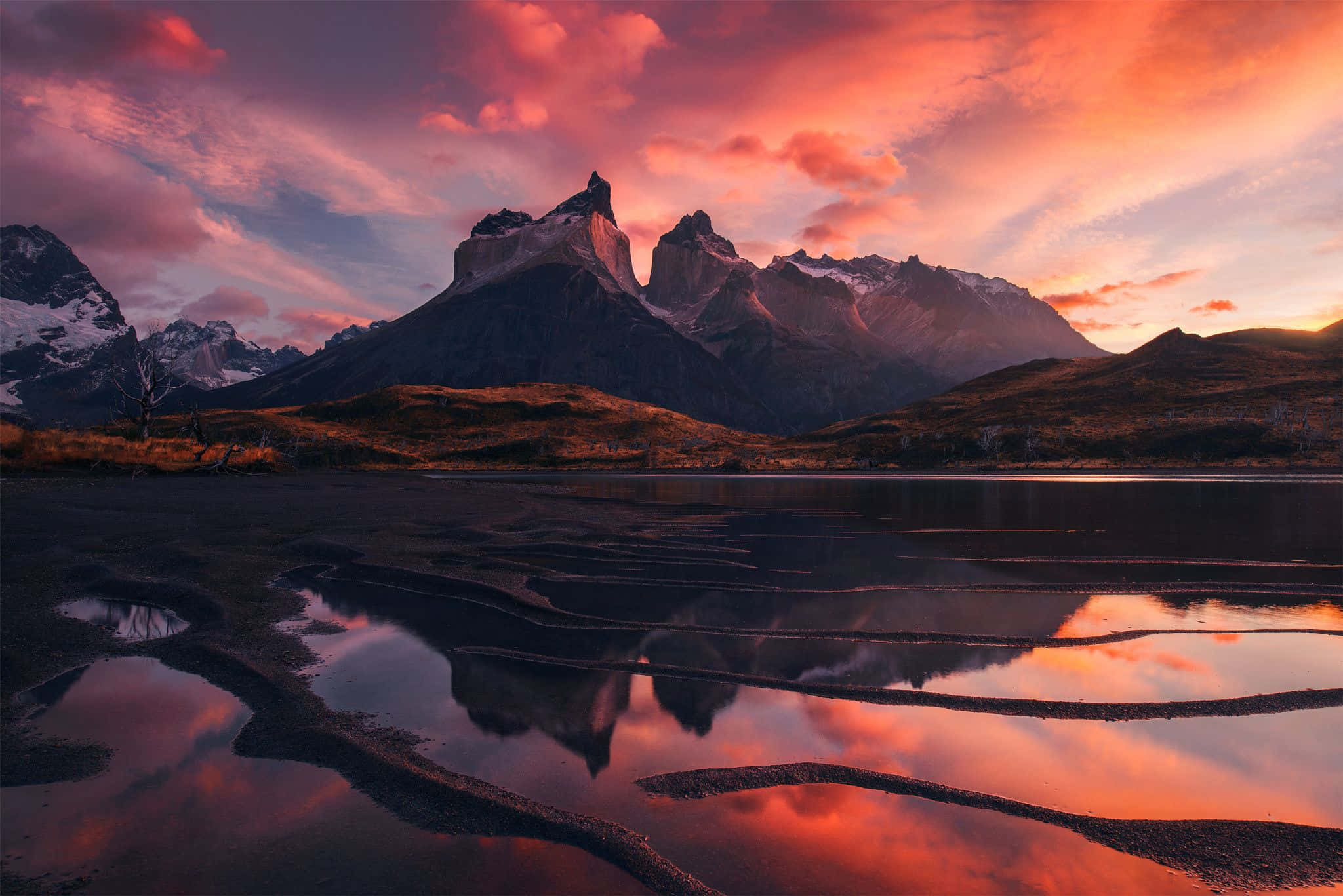 Patagonia 桌布 壁紙 — 一個戲劇性的山脈，白雪覆蓋的山峰在火紅的日落天空下倒映在淺淺的、泛起漣漪的湖面上。