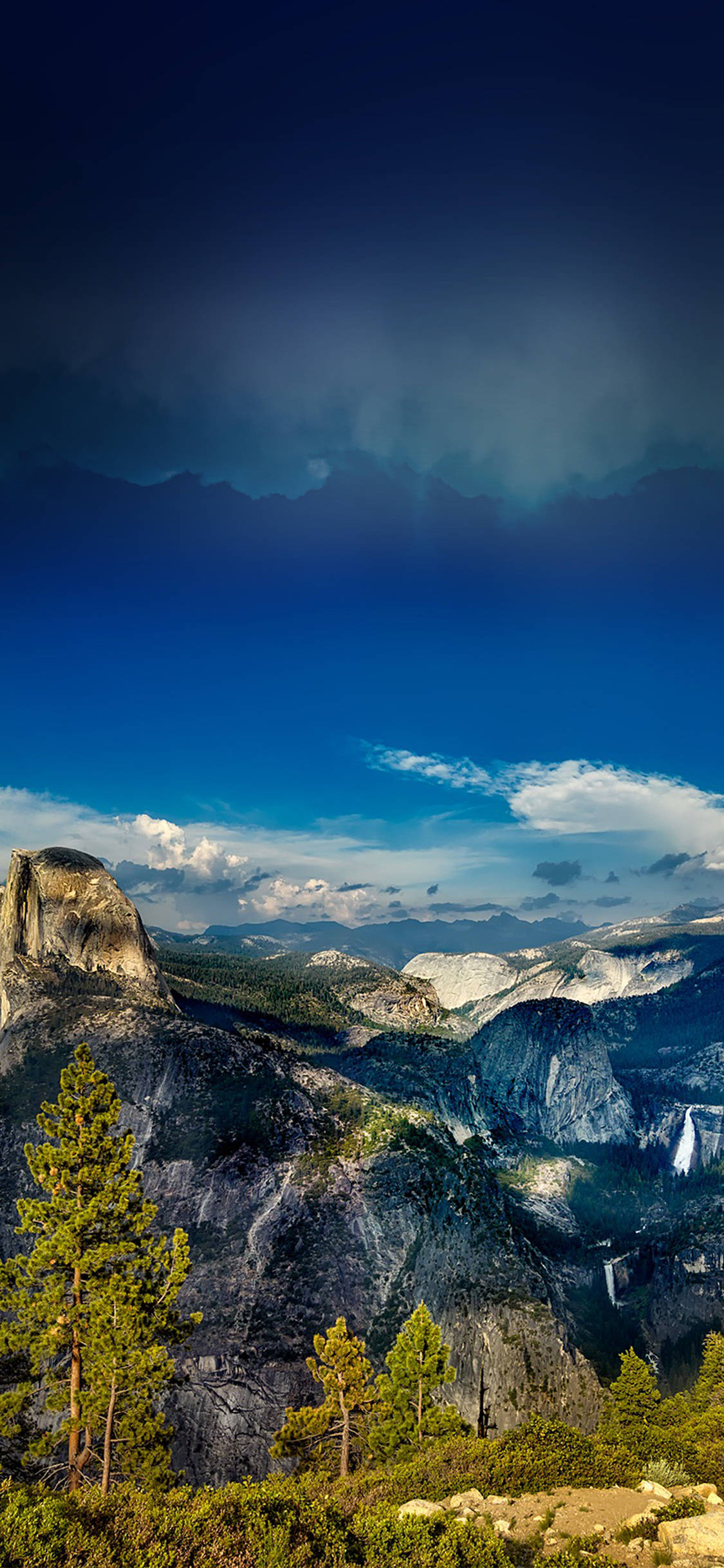 Yosemite iPhone 桌布 壁紙 — 一個戲劇性的山谷，半圓頂在深藍色天空下，散落雲朵和前景的松樹。