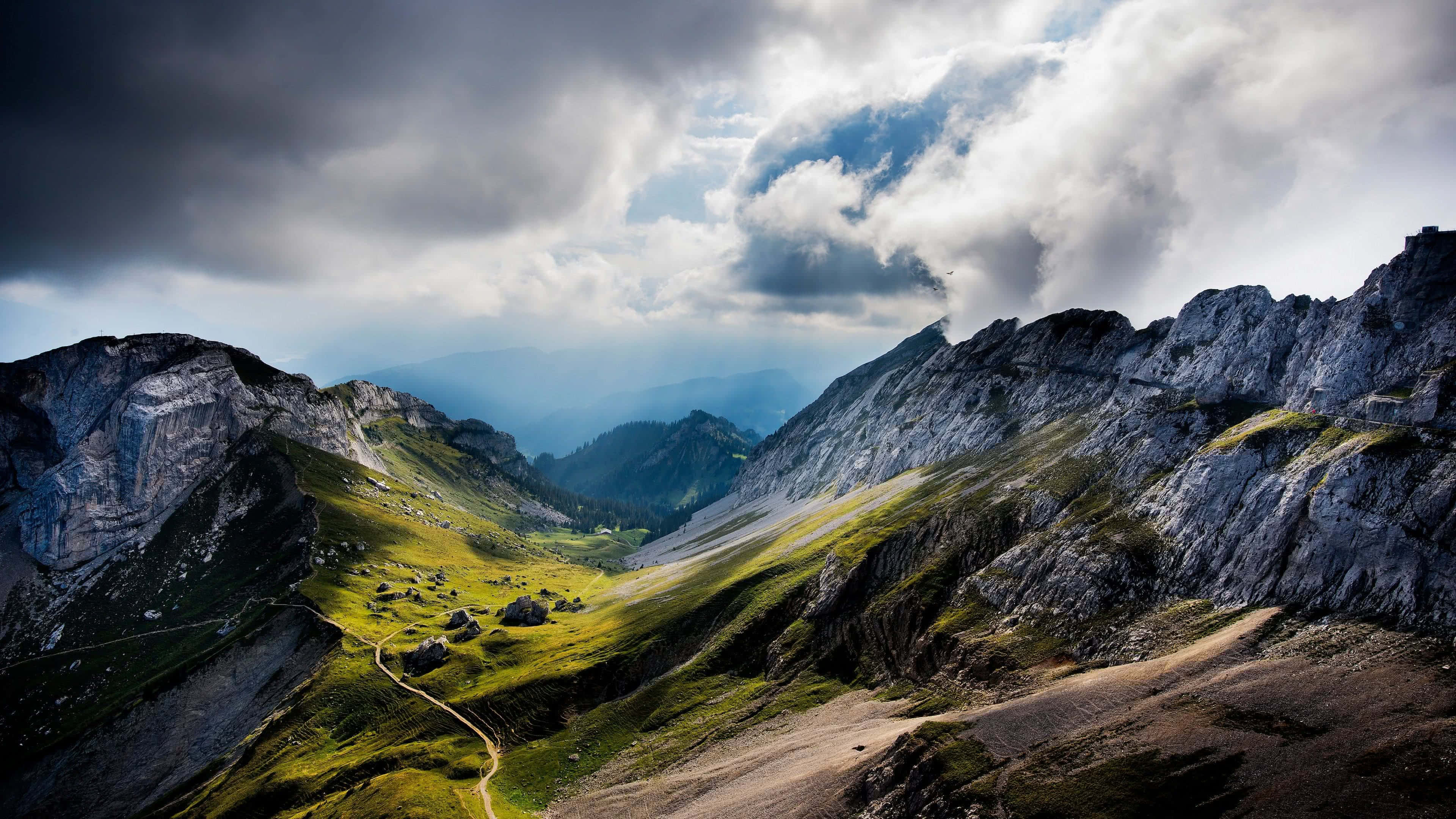 4K Mountain 桌布 壁紙 — 一個戲劇性的山谷，有崎嶇的山峰和蜿蜒的小徑穿過充滿活力的綠色草地，在暴風雨的天空下。